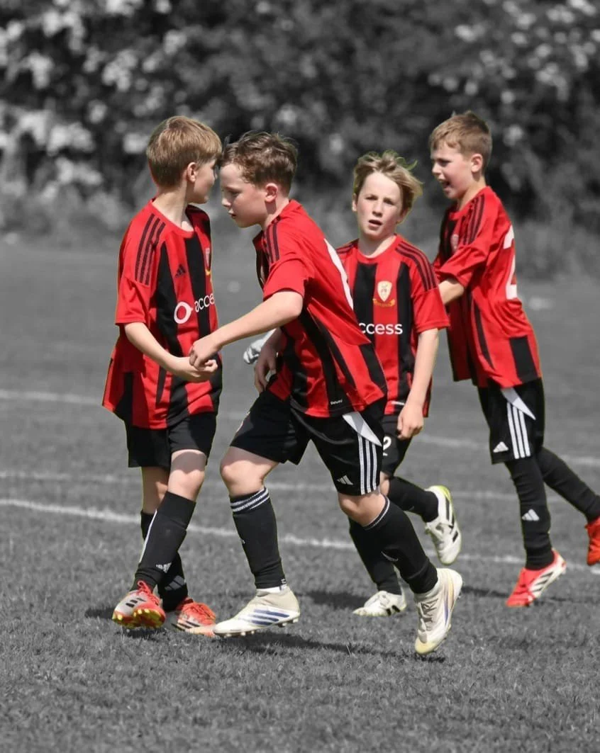 Four young boys in red and black soccer uniforms on a field, two of them are posturing with their hands clasped and the other two are looking on, one with a concerned expression, and the other smiling with his arms crossed.