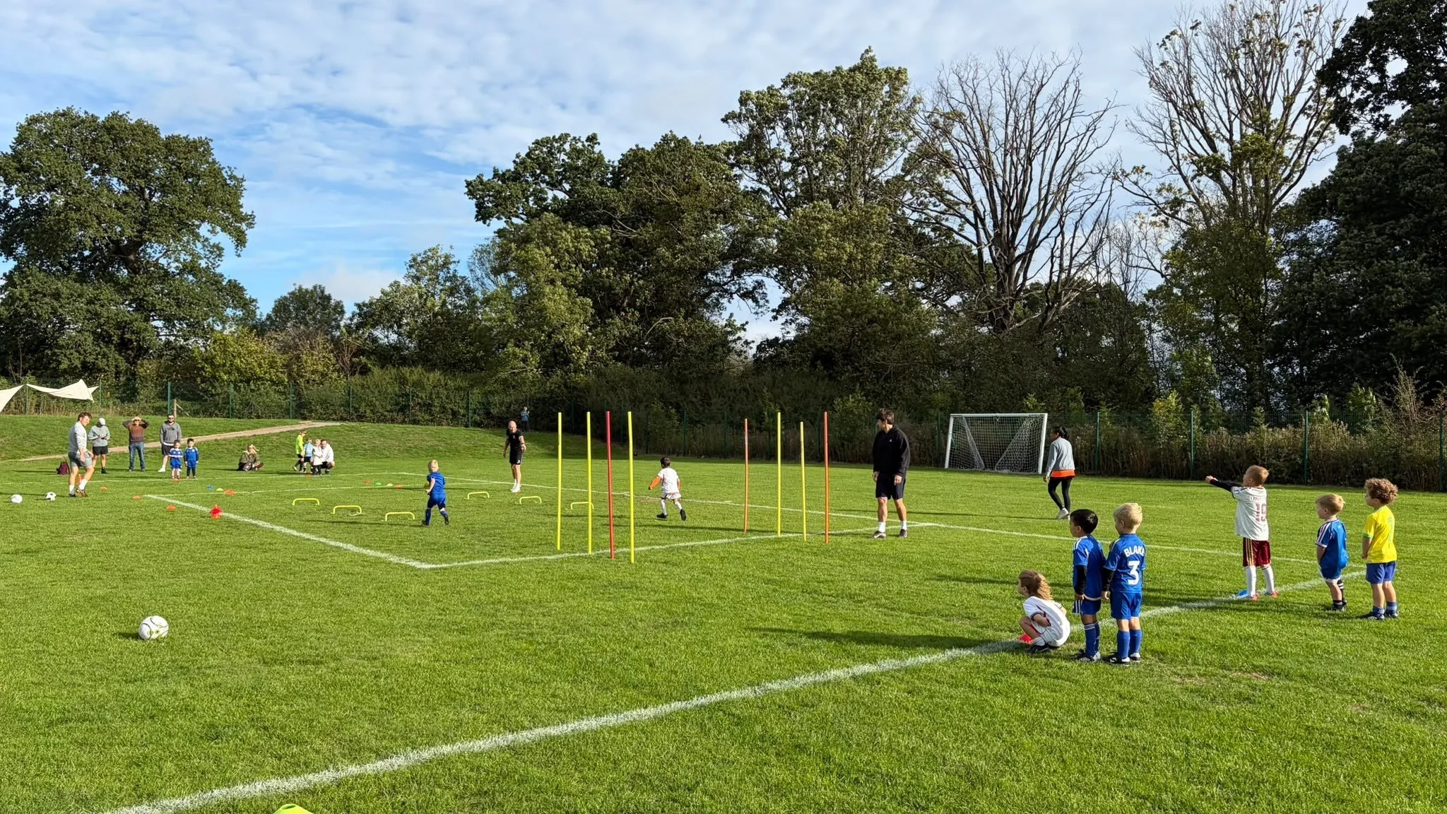 Children playing soccer and practicing drills on a grassy field with coaches, surrounded by tall trees under a partly cloudy sky.