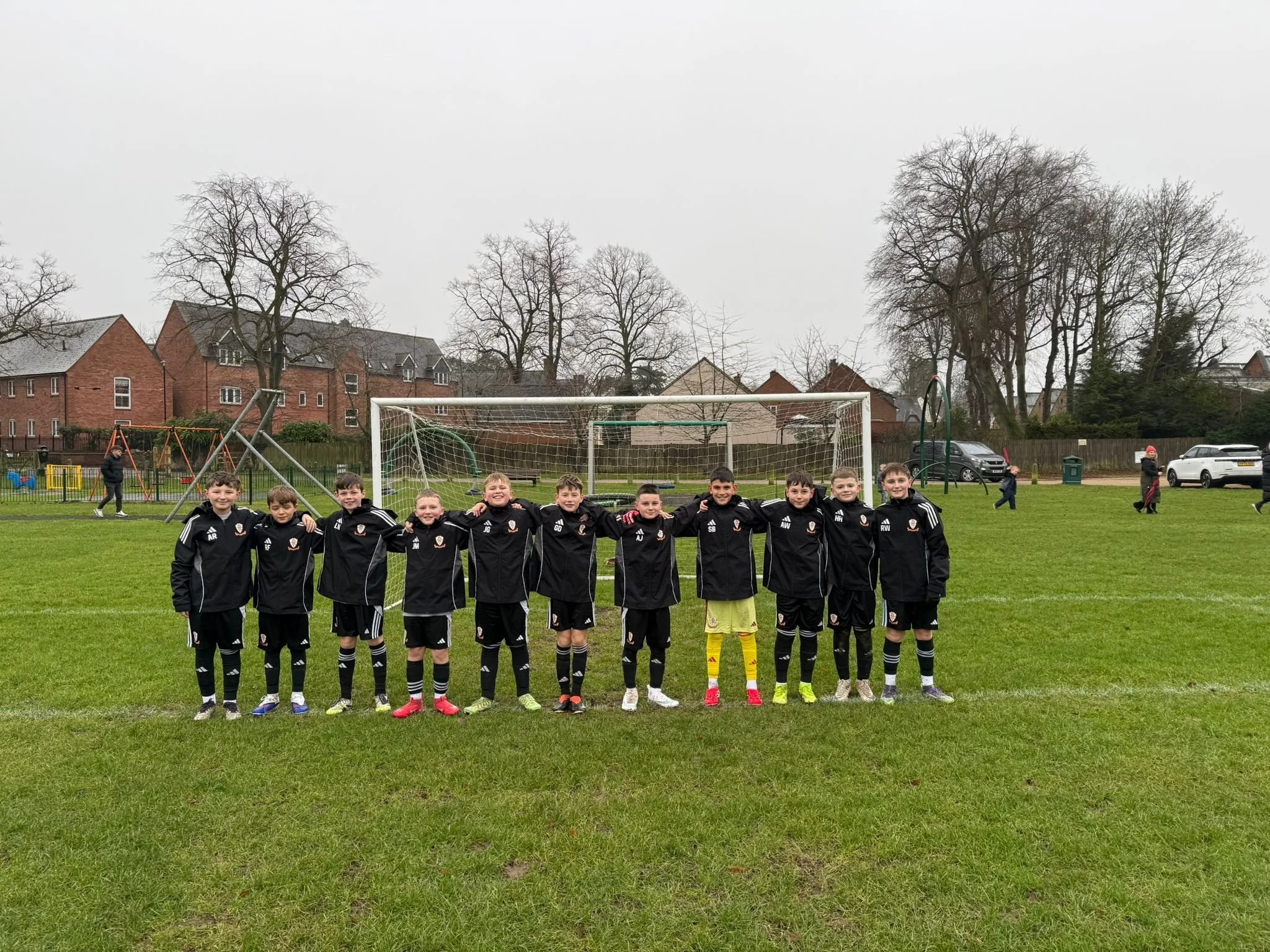 A group of young boys in black soccer uniforms smile and pose together on a soccer field, with a goalpost behind them. There are trees and houses in the background under a cloudy sky.