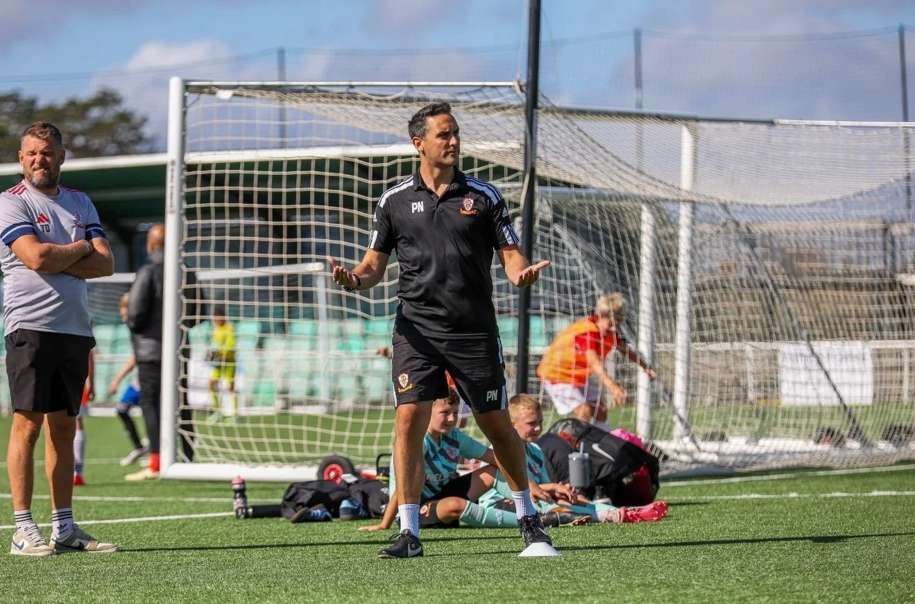 Soccer coach gesturing during a practice session on a sunny day, with players sitting on the ground and a goalpost in the background.