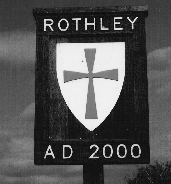 A wooden sign with the name "Rothley" at the top, a shield with a cross in the center, and the text "AD 2000" at the bottom.