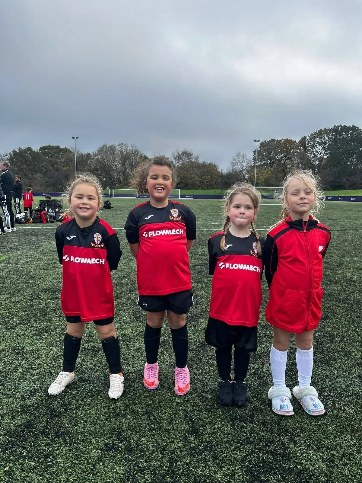 Four young girls standing on a soccer field, wearing black and red soccer jerseys with the logo 'FLOWMECH,' smiling and posing for a photo, with other players and coaches in the background under a cloudy sky.