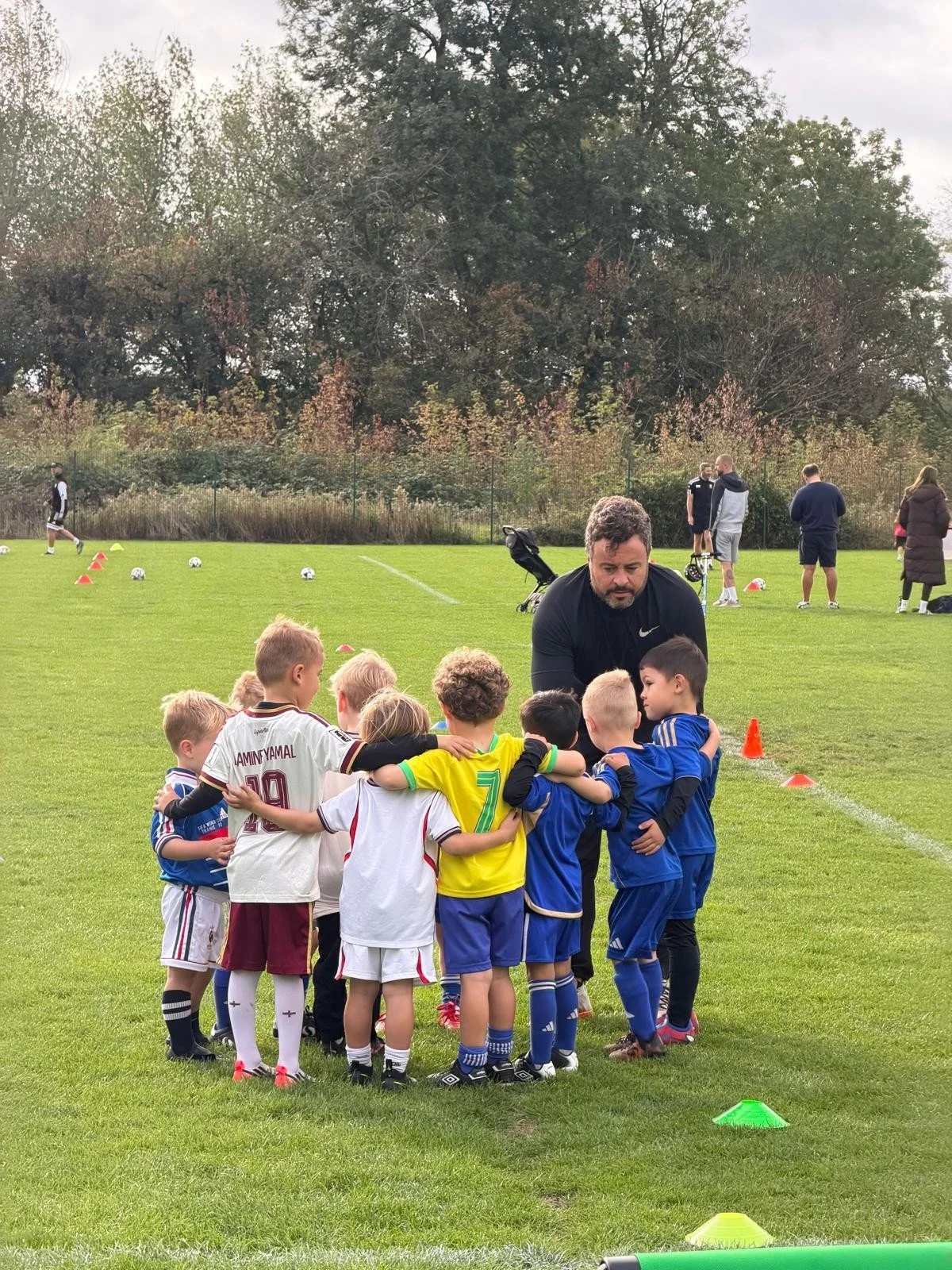 Young children dressed in soccer uniforms gather around their coach for a team huddle on a soccer field during practice, with other players and coaches in the background.