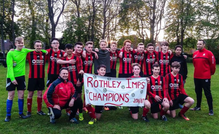 A youth soccer team posing outdoors on a grassy field, holding a sign that says 'Rothey Imps Champions 2016/17'. The team members are wearing red and black striped jerseys, with two coaches standing on either side. The sun is setting in the background, and trees are visible.