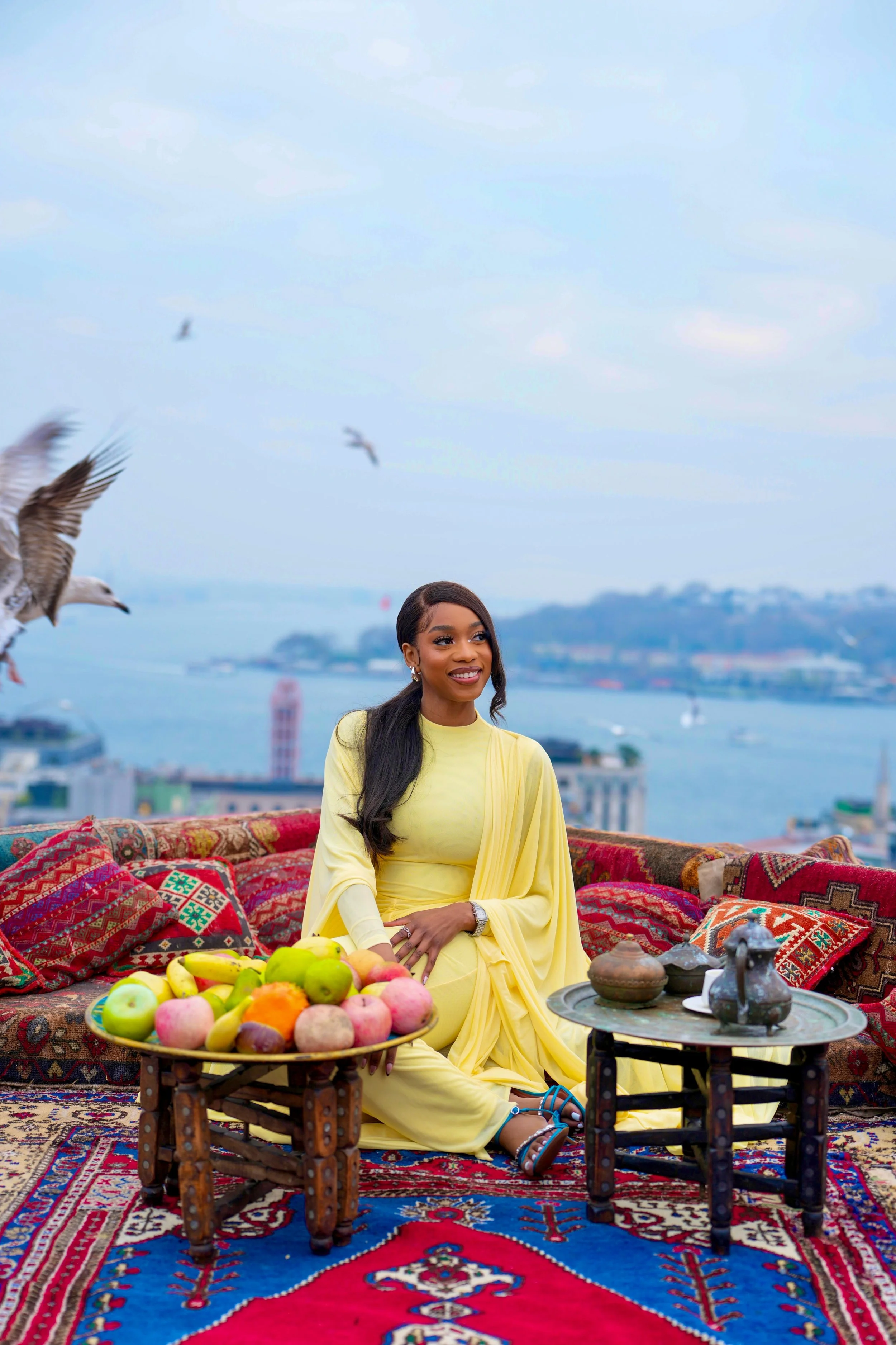 A woman dressed in yellow attire is sitting on a colorful traditional rug on a rooftop during daytime, surrounded by an assortment of fruits, including apples and bananas, with a cityscape and waterway in the background. There are birds flying in the sky and decorative cushions and a small table with traditional items nearby.