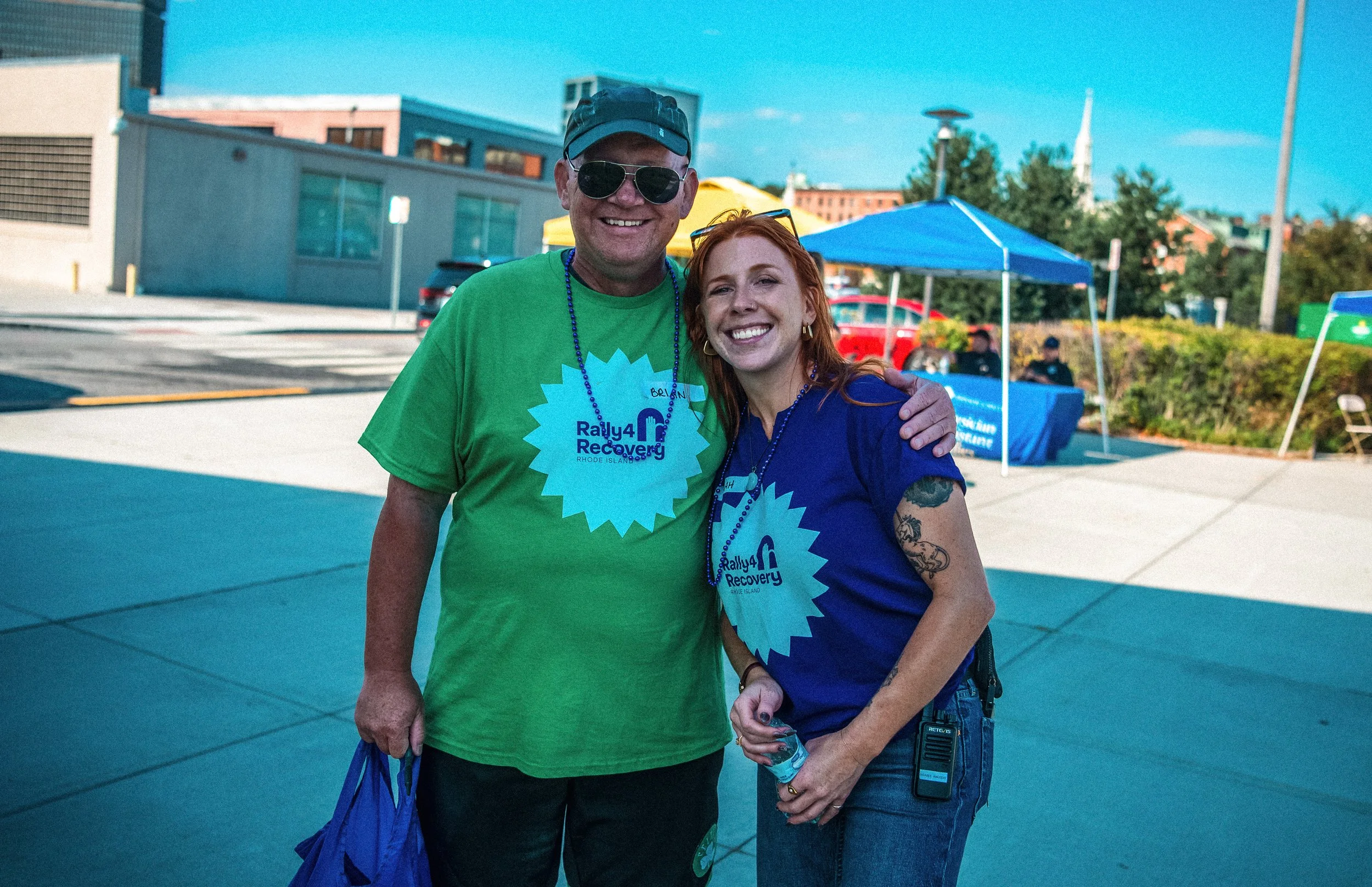 A smiling man and woman at an outdoor event, wearing matching blue and green T-shirts with a 'Rally4Recovery' logo, standing on a paved area with colorful tents and trees in the background.