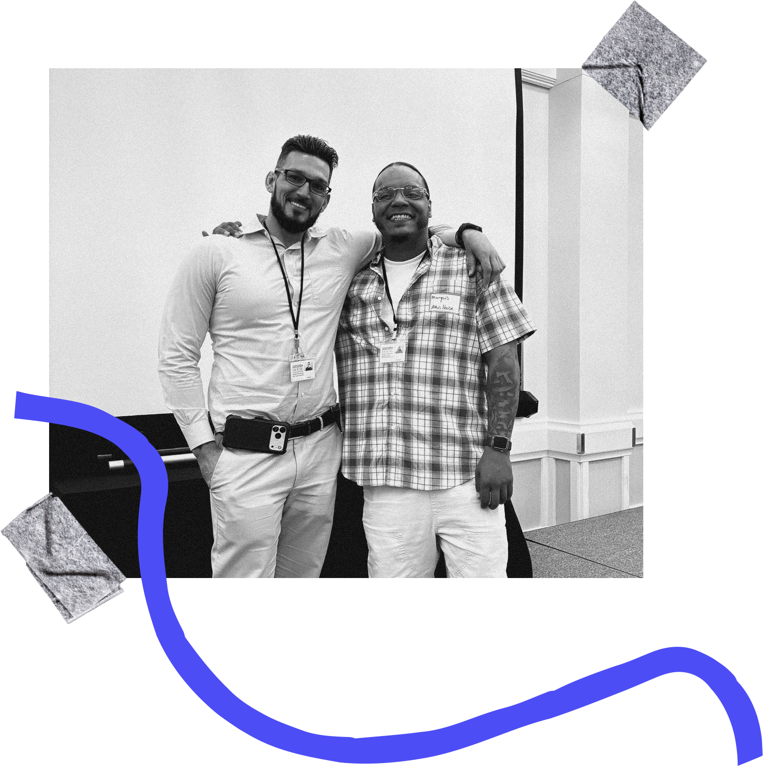 Two men standing together, smiling, with one having his arm around the other, in an indoor setting with a white wall background.