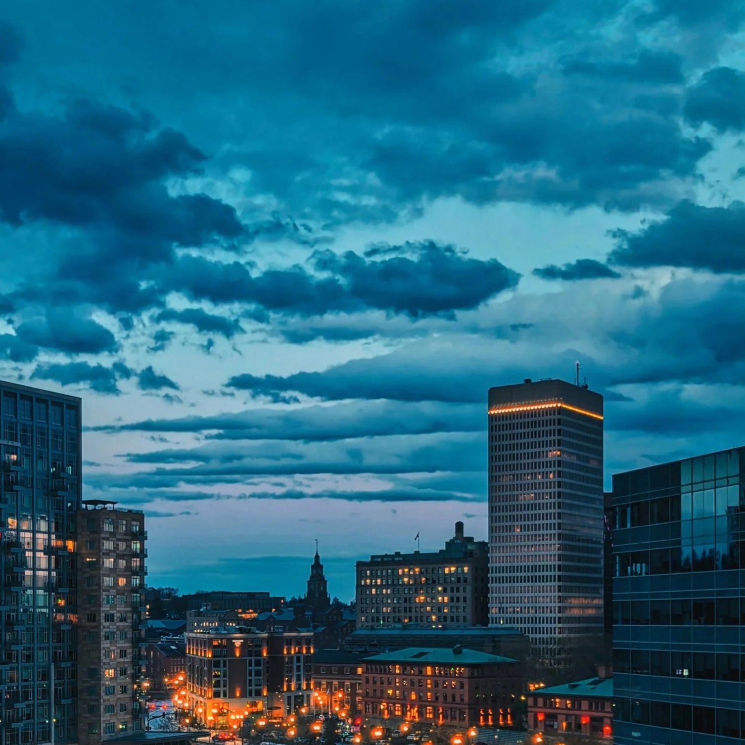 City skyline at dusk with tall buildings, some with illuminated windows, and a cloudy sky overhead.