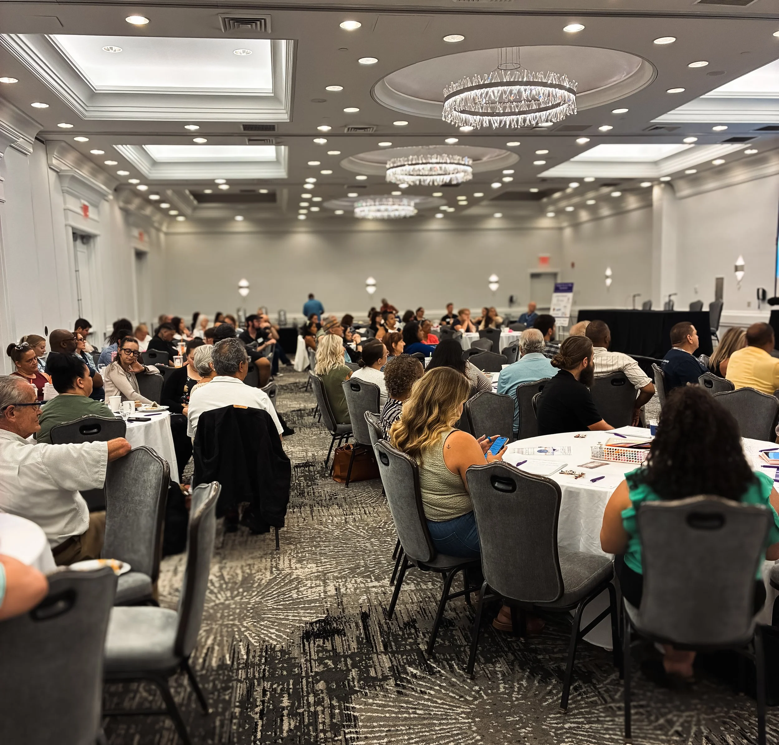 Conference room filled with seated attendees, large chandeliers hanging from a decorated ceiling, and patterned carpet.