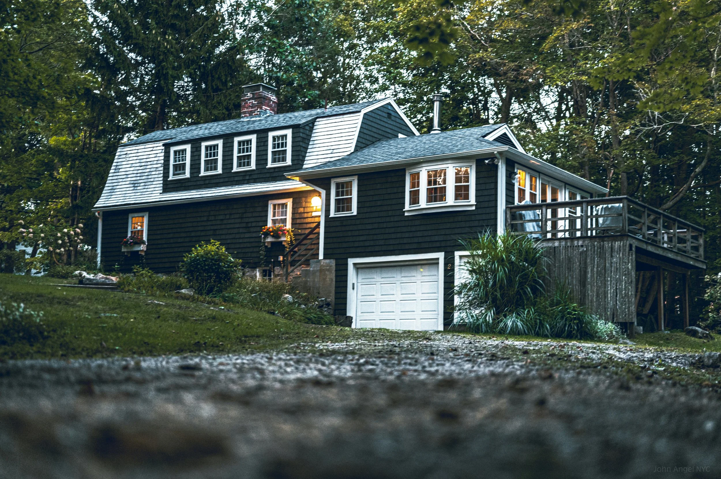 A house with black exterior siding, multiple windows with white frames, and a driveway with a garage door at the front. The house has a chimney, a deck to the right, and is surrounded by lush trees and greenery.