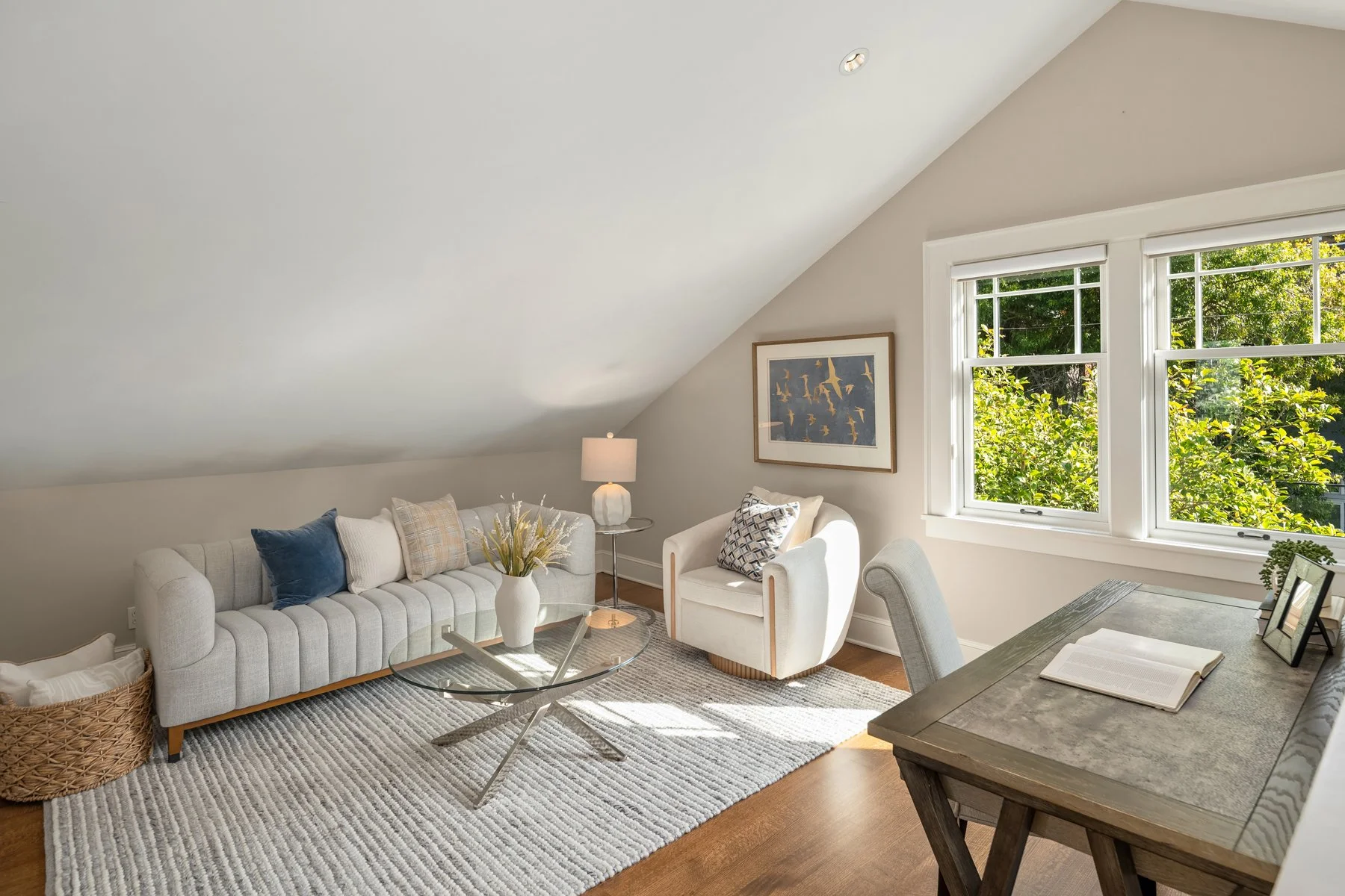 Living room with white sofa, armchair, glass coffee table, side table with lamp, wall art, large window with greenery outside, and a nearby dining table with open book.