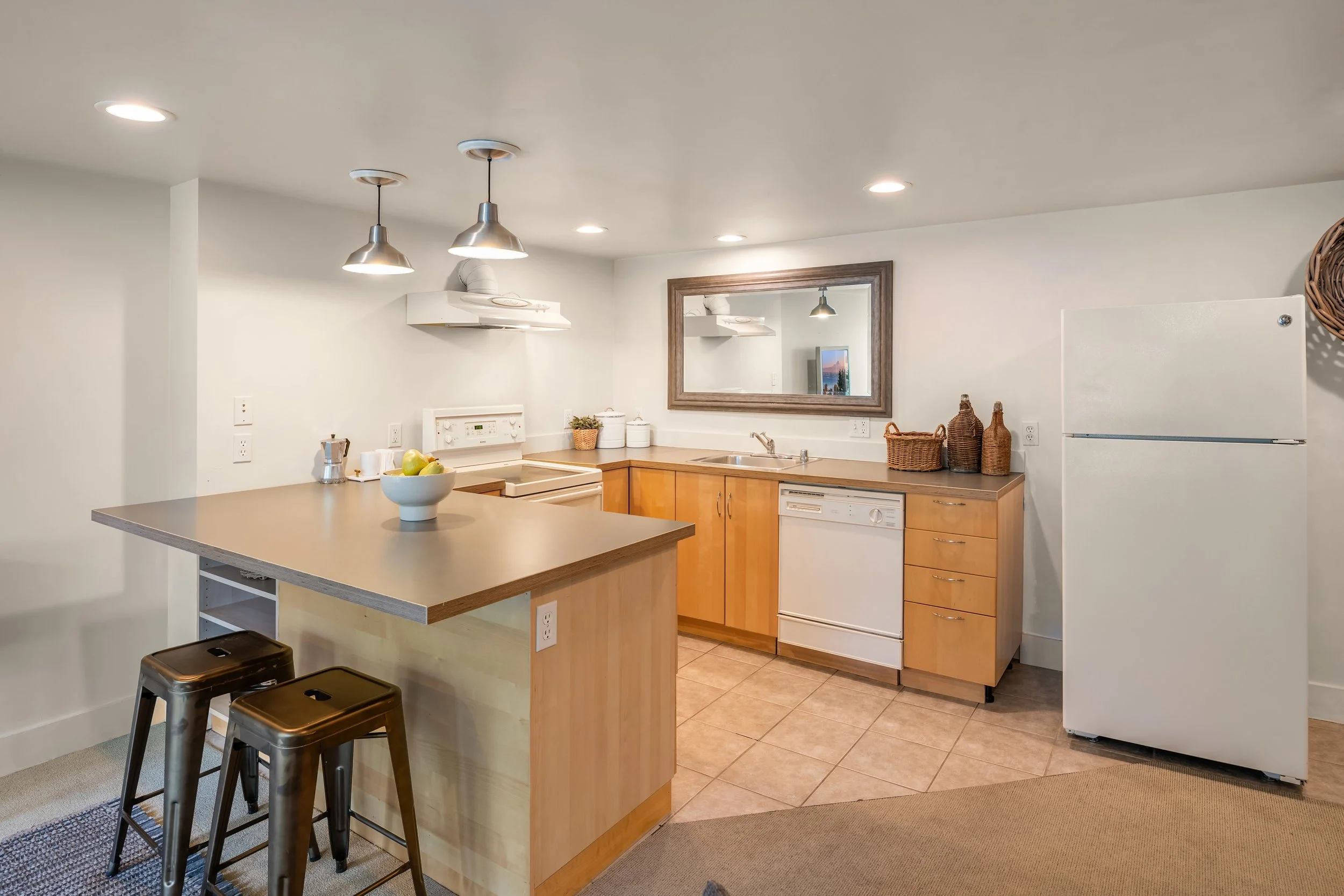 Kitchen with light wood cabinets, white appliances, and a beige countertop. Items include a white bowl of green apples, decorative baskets, and a mirror on the wall. Overhead lighting and a ceiling exhaust are visible.