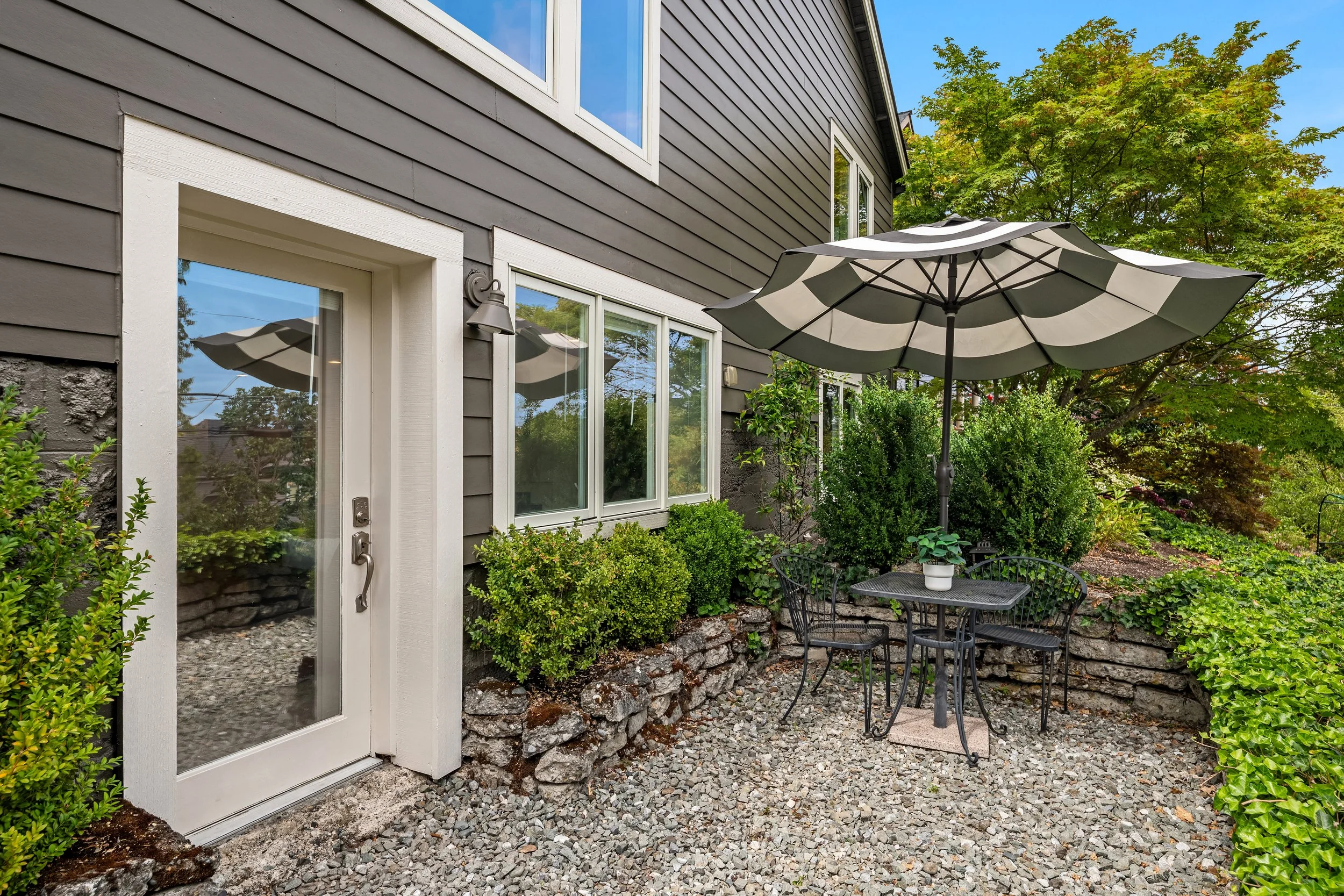 A backyard patio with black metal table and chairs, a large striped umbrella, surrounded by green bushes and plants, and the exterior of a gray house with white trim and large windows.