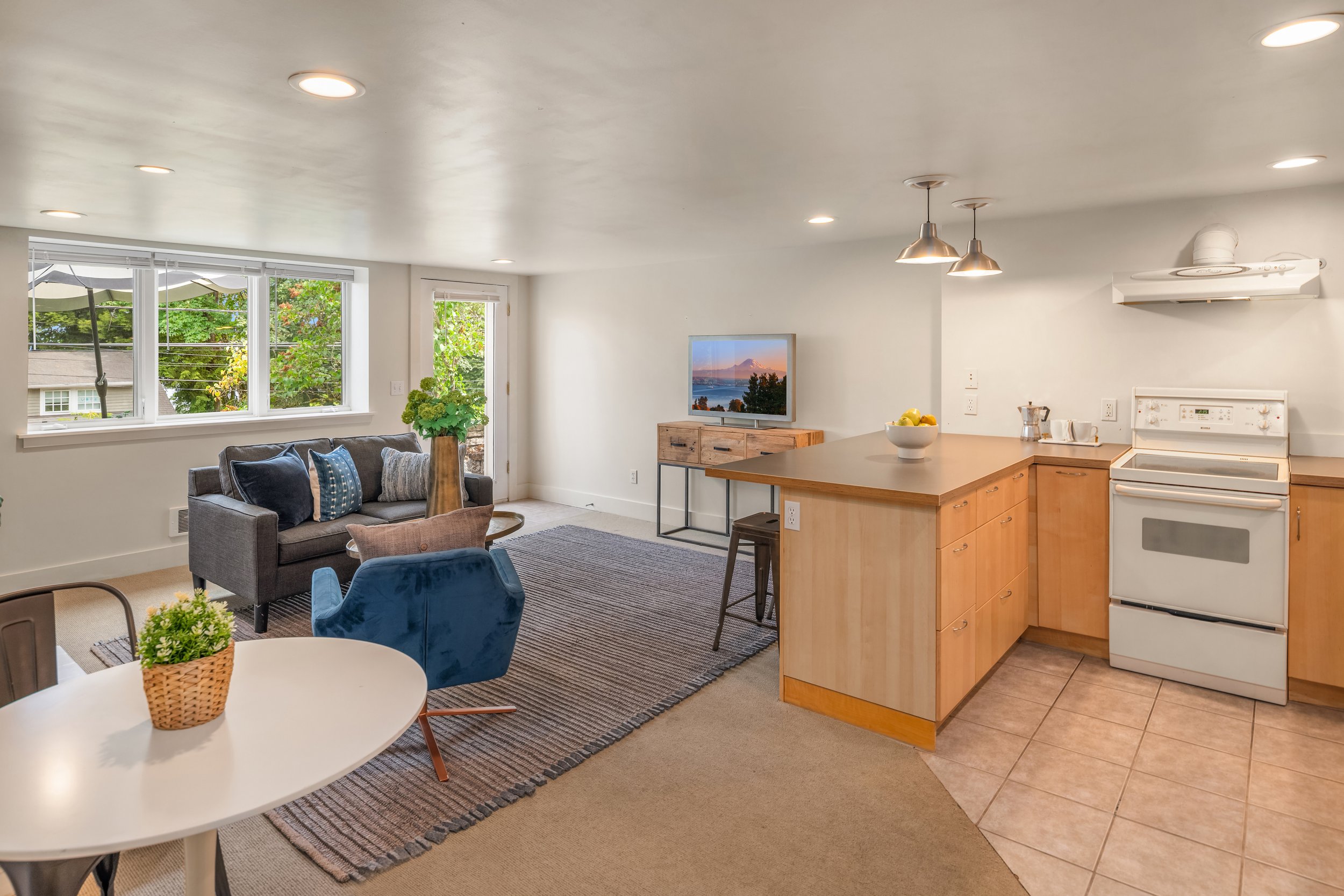 Open-concept living room and kitchen with large window, gray sofa, dining table with plants, and a kitchen counter with fruit bowl, stove, and pendant lights.