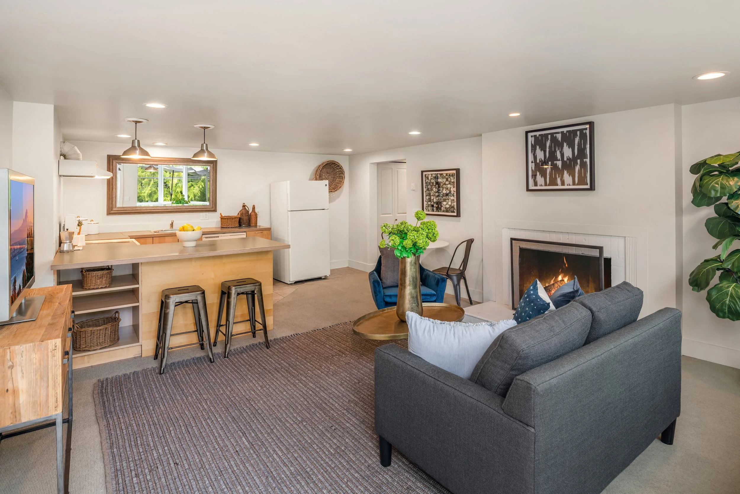 Living room with gray sofa, fireplace, artwork, green plant, and dining area with table and chairs, in a modern home.