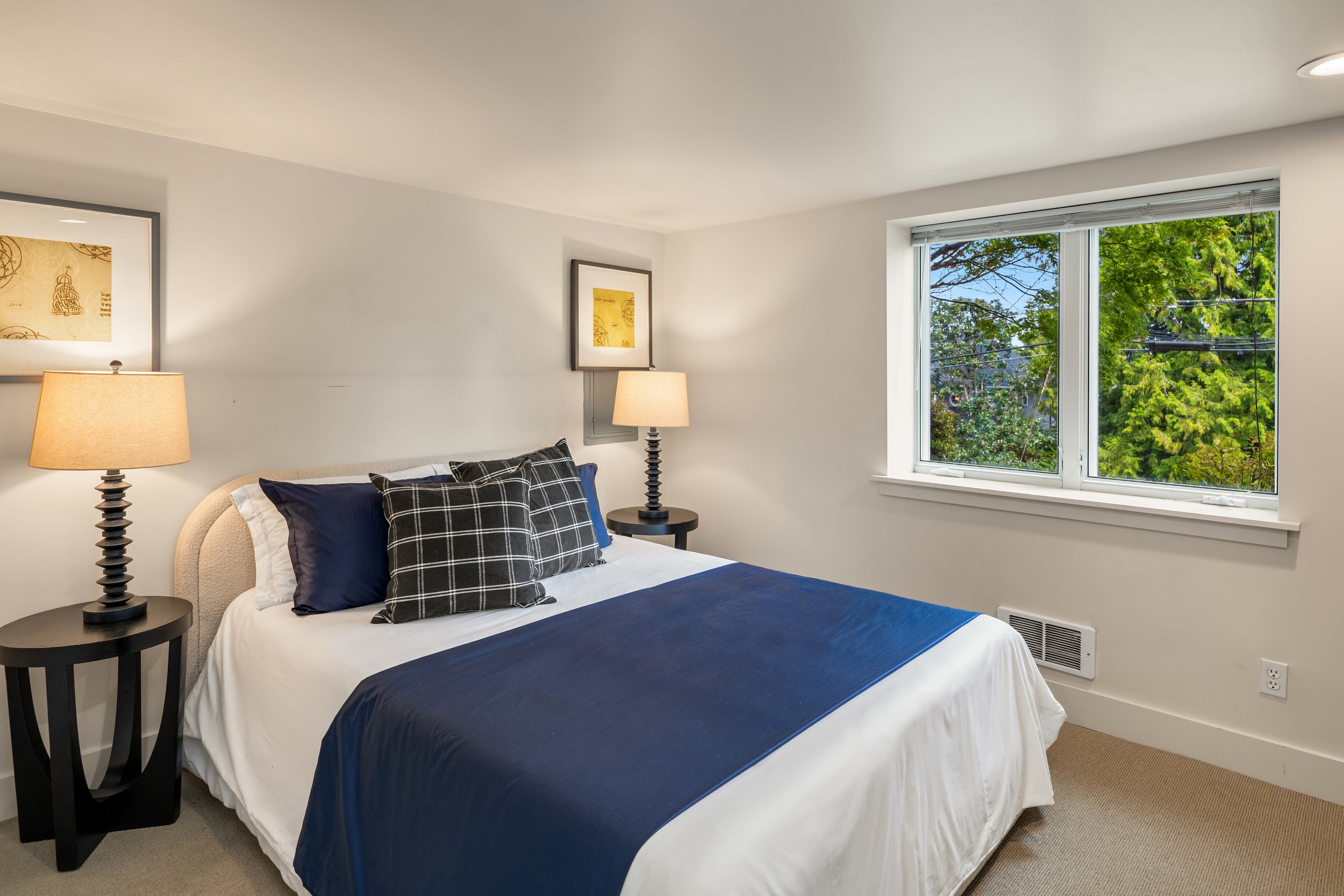 A bedroom featuring a bed with black, white, and navy blue bedding, two pillows, and a window showing green trees outside. There are two black nightstands with lamps and framed artwork on the wall.