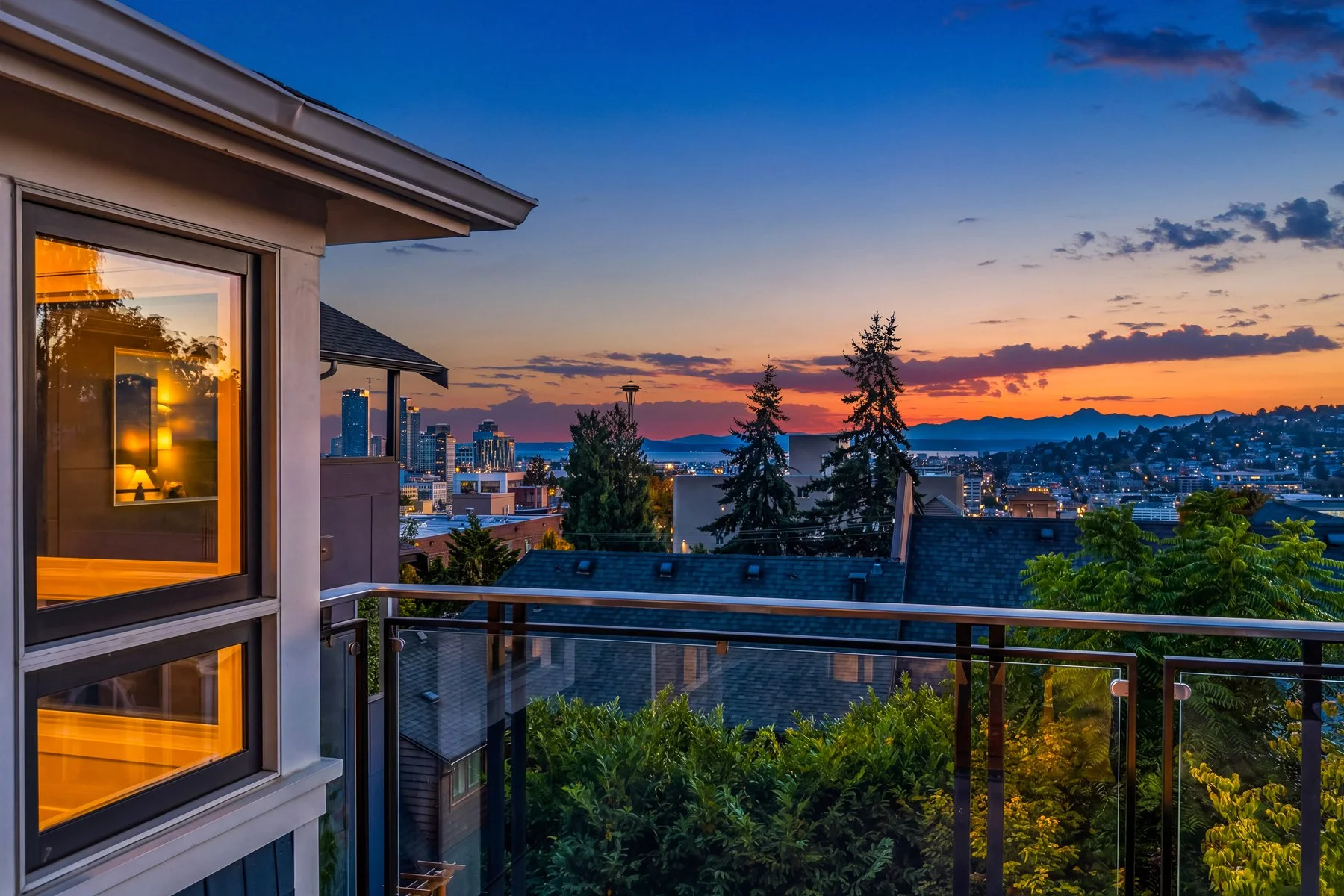 A balcony view overlooking a city skyline at sunset, with trees and rooftops in the foreground and tall buildings illuminated in the distance.