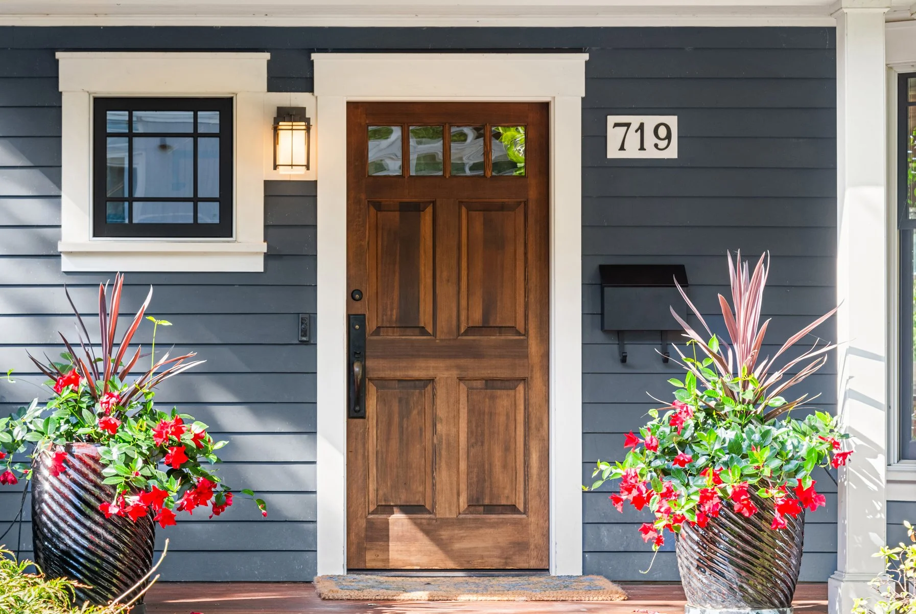 A front door with a dark wood finish and glass window panels at the top, surrounded by white trim, with a black house number sign displaying 719 to the right and flowers in two large striped planters on either side, against a gray horizontal siding e