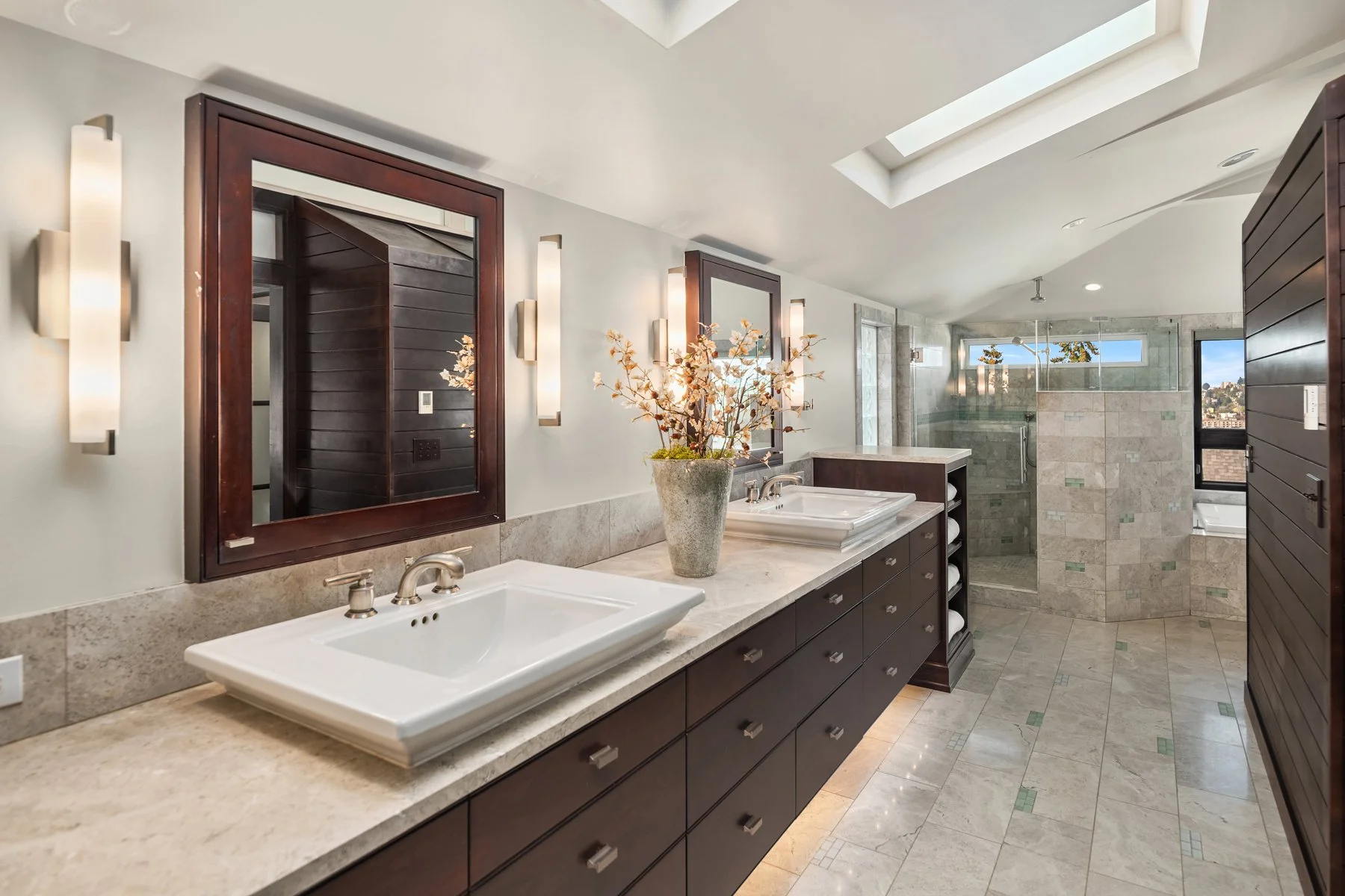 Modern bathroom with double sinks, large mirrors, a skylight, a window, and a spacious shower and bathtub area, decorated with a vase of flowers.