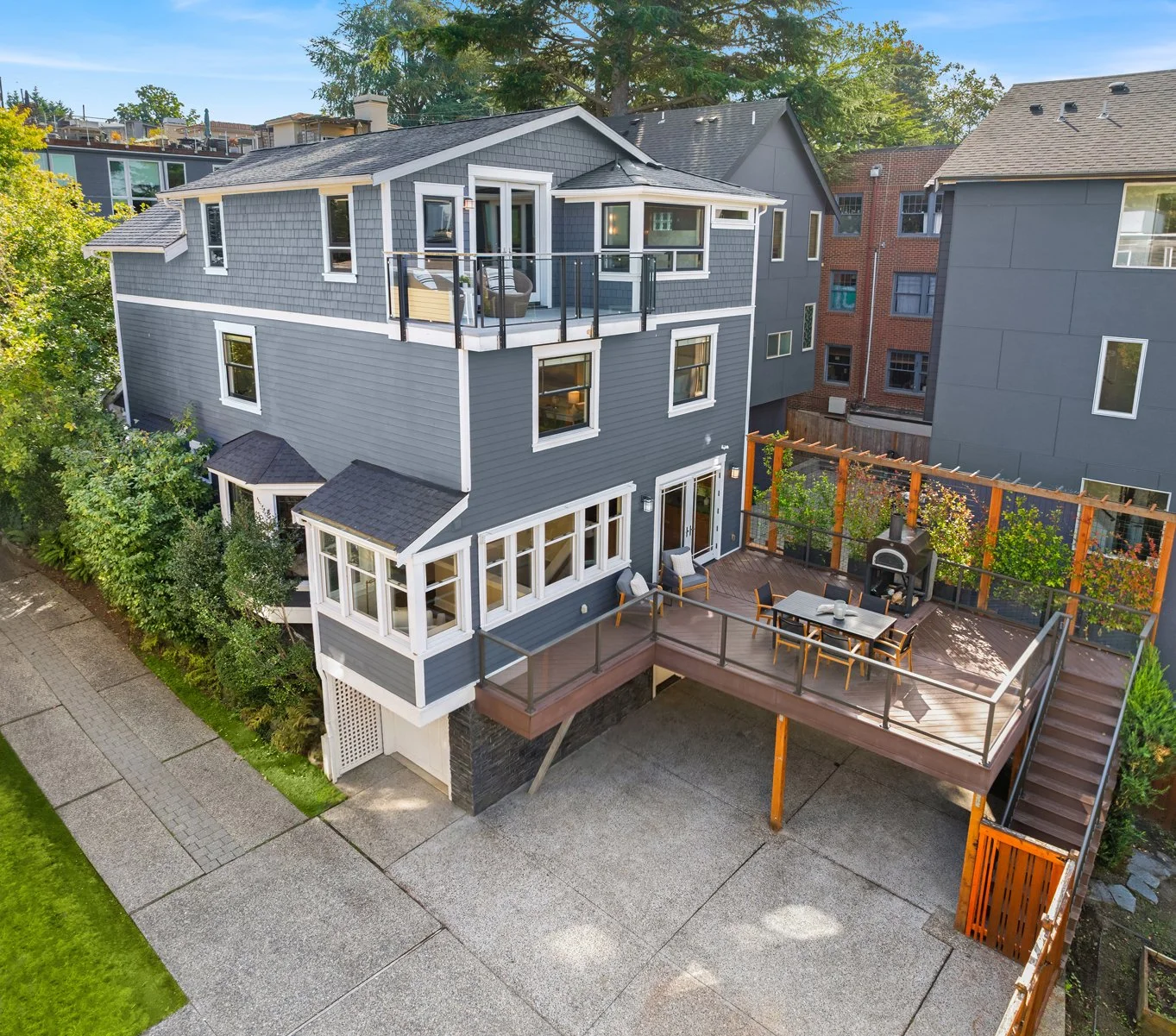 A modern, multi-story house with gray siding and large windows, featuring a spacious wooden deck with outdoor furniture, a pizza oven, and surrounding greenery.