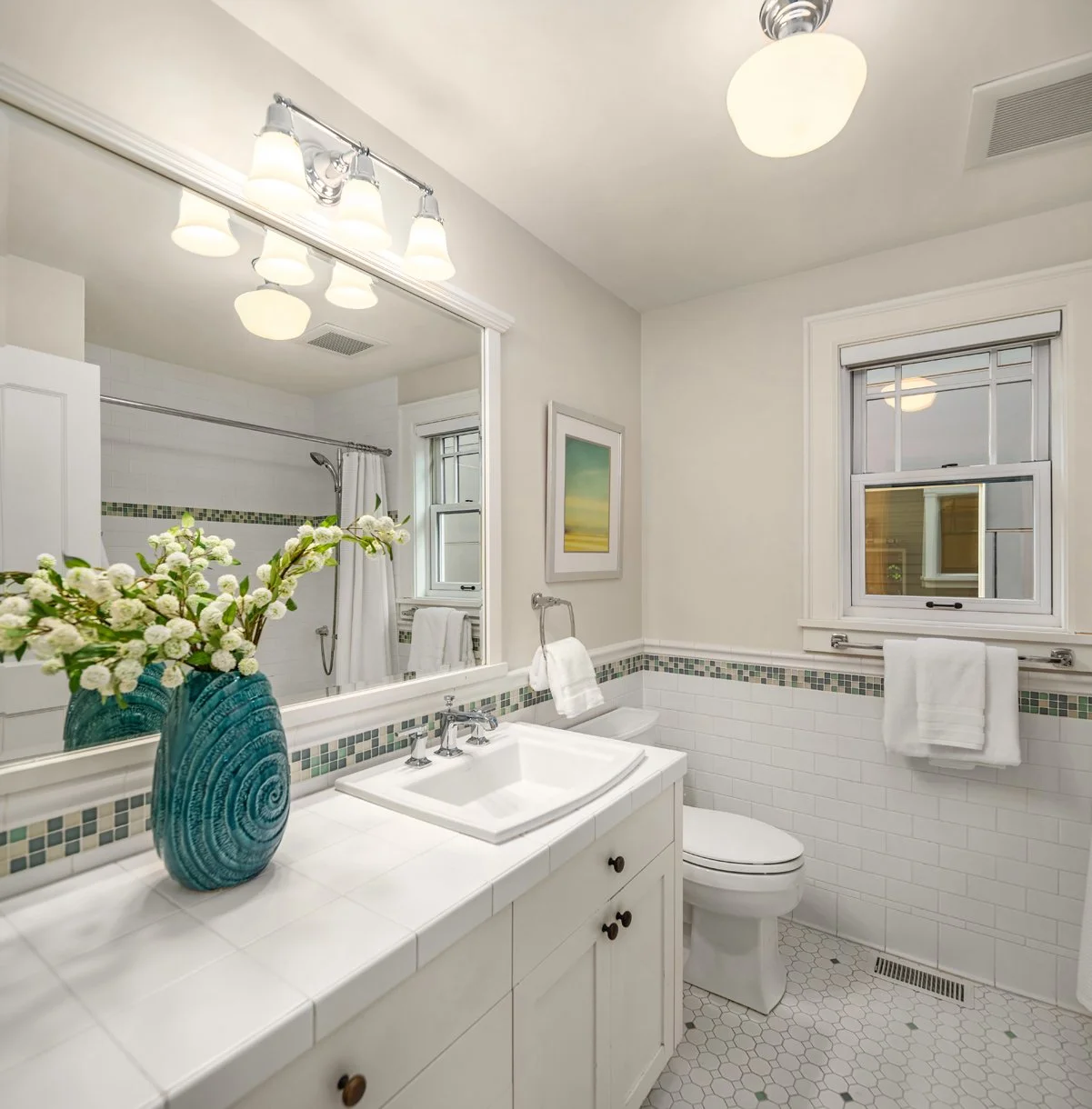 Bright bathroom with white tile walls and floor, featuring a white vanity with a sink, a large mirror, a toilet, two windows, and decorative elements including a blue vase with white flowers and a landscape painting.