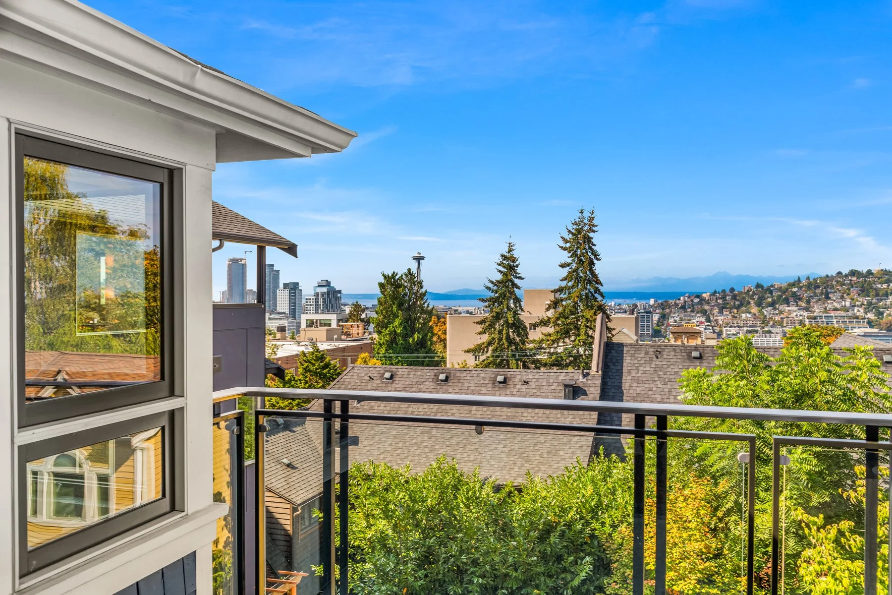A city view from a balcony with trees, rooftops, and skyline of Seattle in the distance under a blue sky.