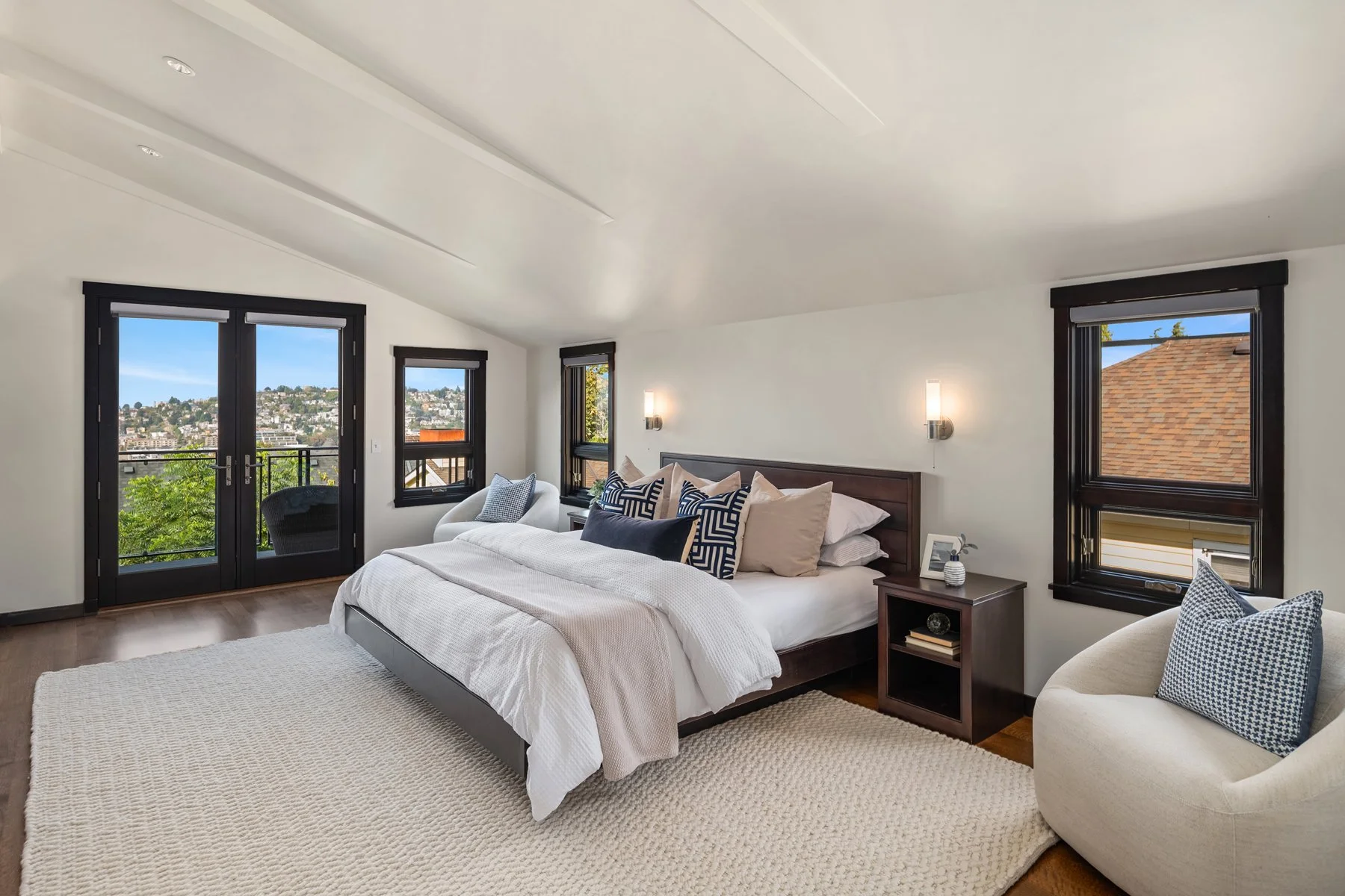 A spacious bedroom with a bed covered in white linens and multiple decorative pillows featuring navy and white patterns. There are three windows with black frames, showing a cityscape and a roof outside. A small bedside table has a framed photo and a