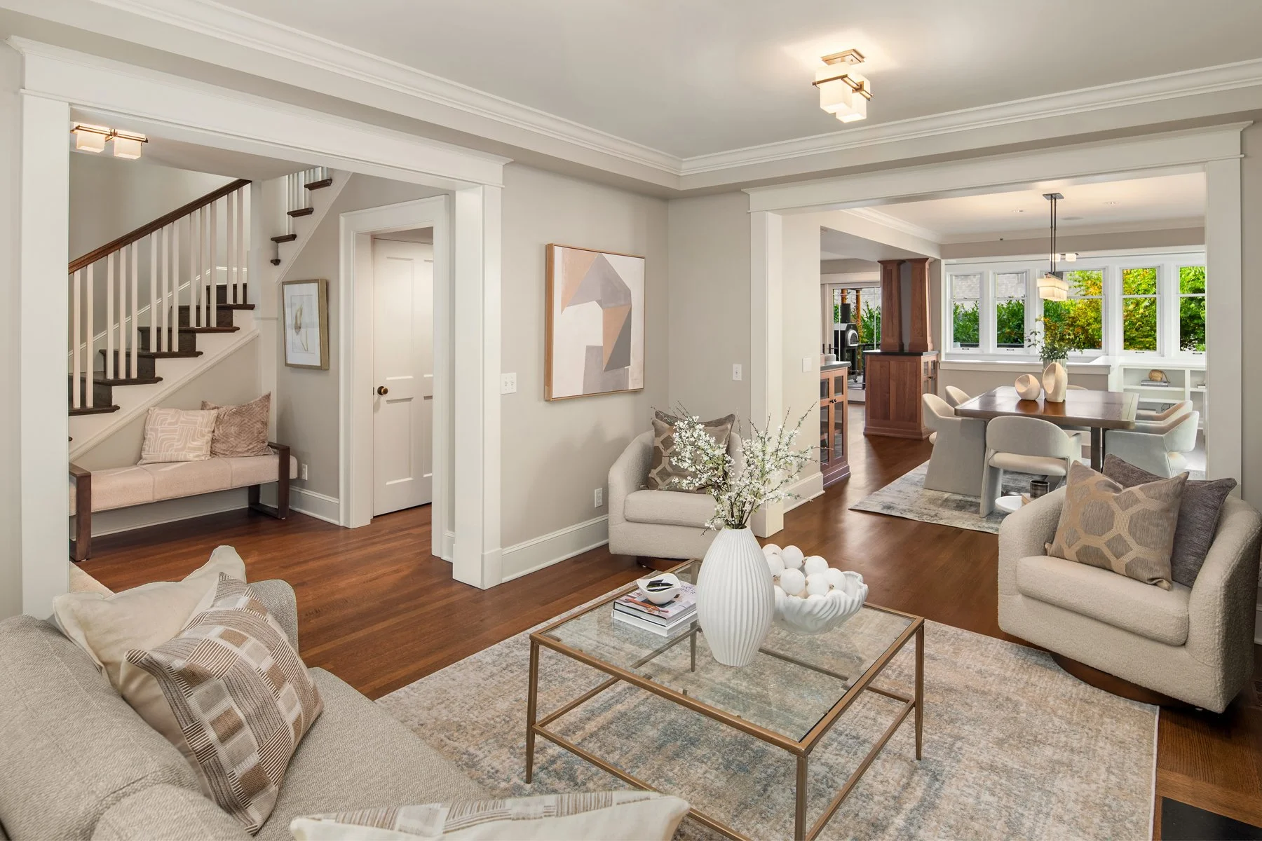 Living room with beige sofa, armchair, and coffee table with white vase and decorative objects; staircase with dark wood steps and white railing; dining area with large windows, wooden table, and white chairs; art pieces on walls; hardwood floors; mo