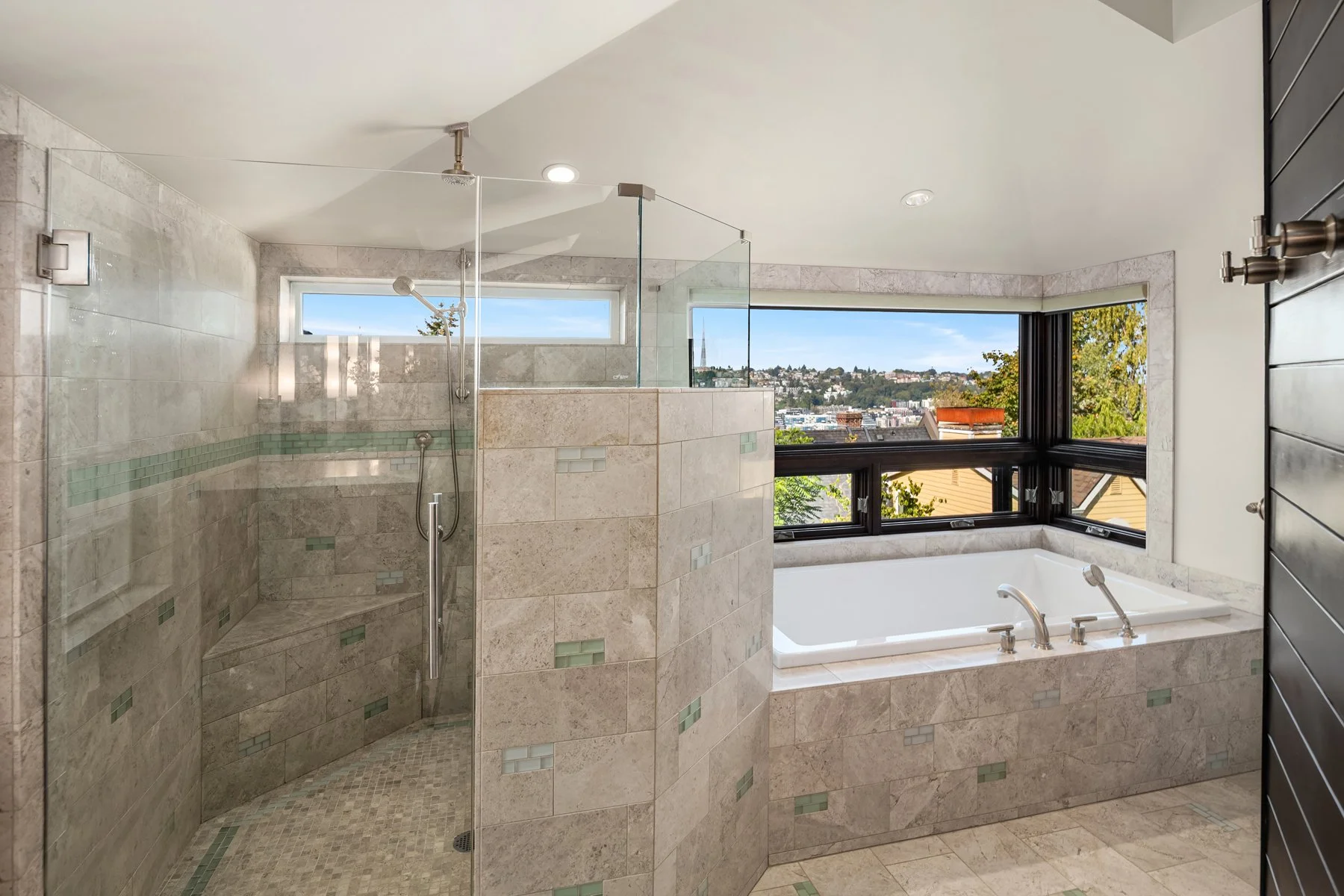 Modern bathroom with a glass-enclosed shower, a bathtub, large windows with a city view, and beige tiled walls and floor.