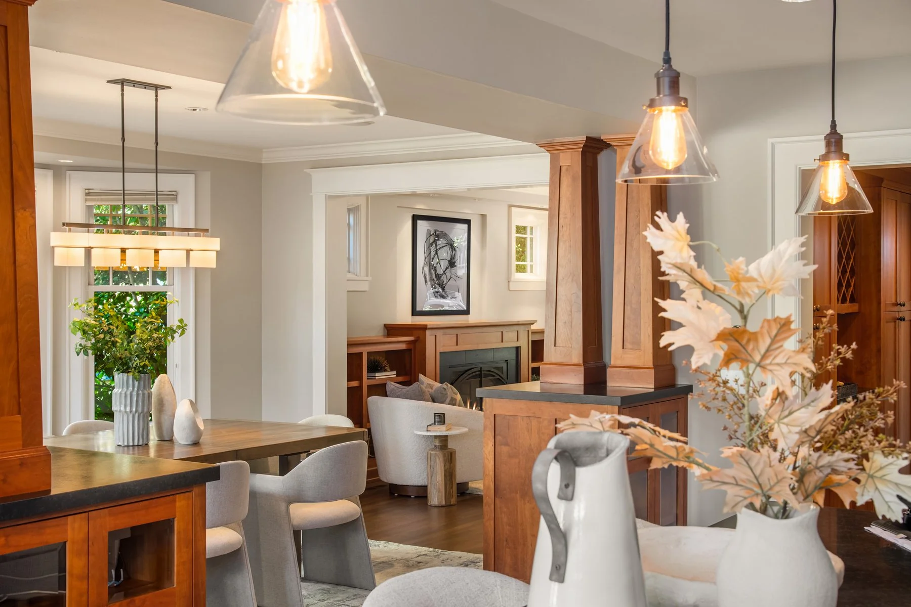 A modern living and dining room with wooden accents, a dining table with beige upholstered chairs, a fireplace, and decorative plants and artwork.