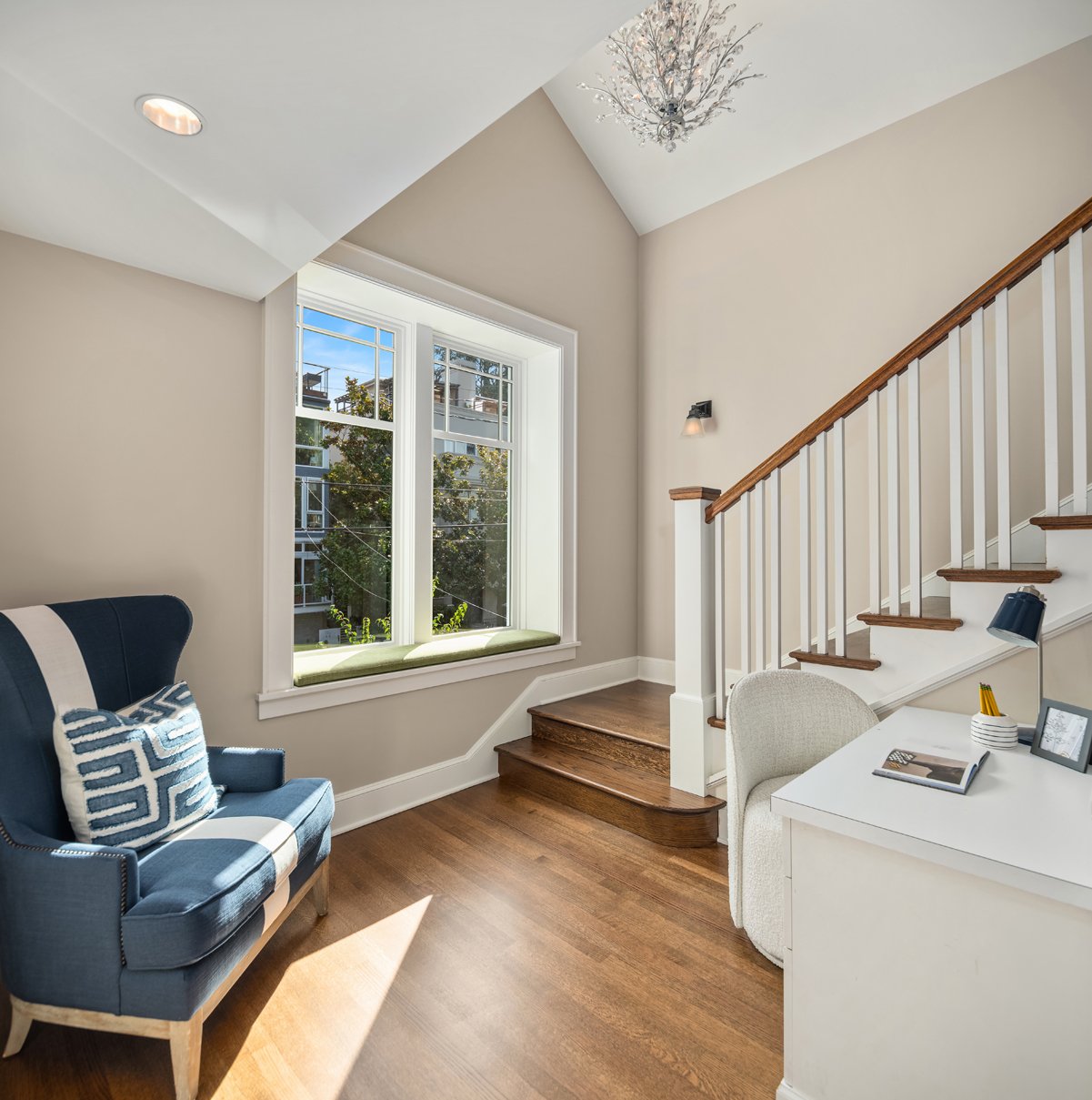 Bright corner of a room with a large window, wooden floors, a staircase with a white railing, a blue armchair with decorative pillows, a white desk with a chair, a lamp, and ceiling light fixtures.