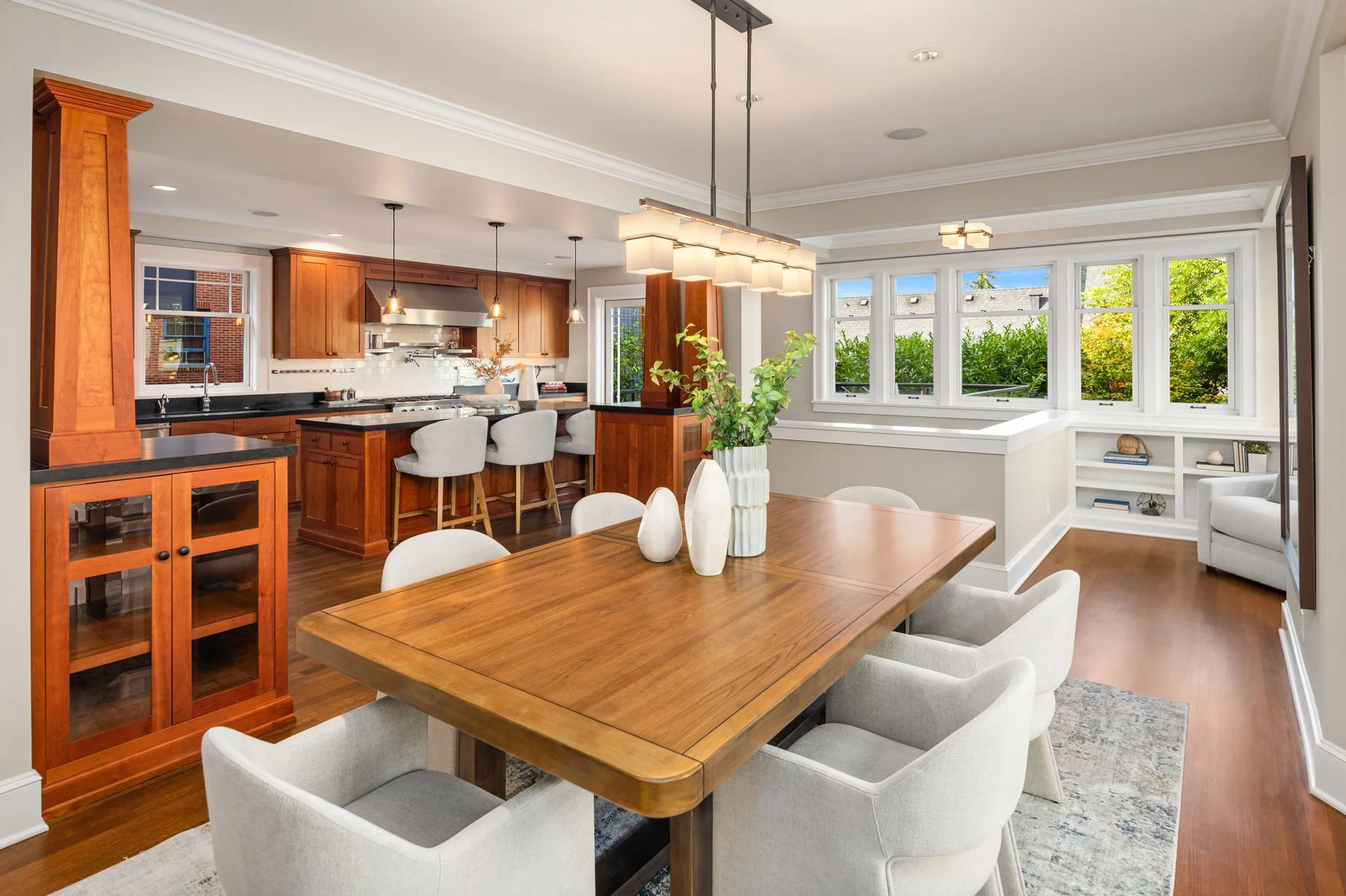 Modern open-concept kitchen and dining area with wooden cabinetry, a dining table with six chairs, large windows, and decorative vases on the table.