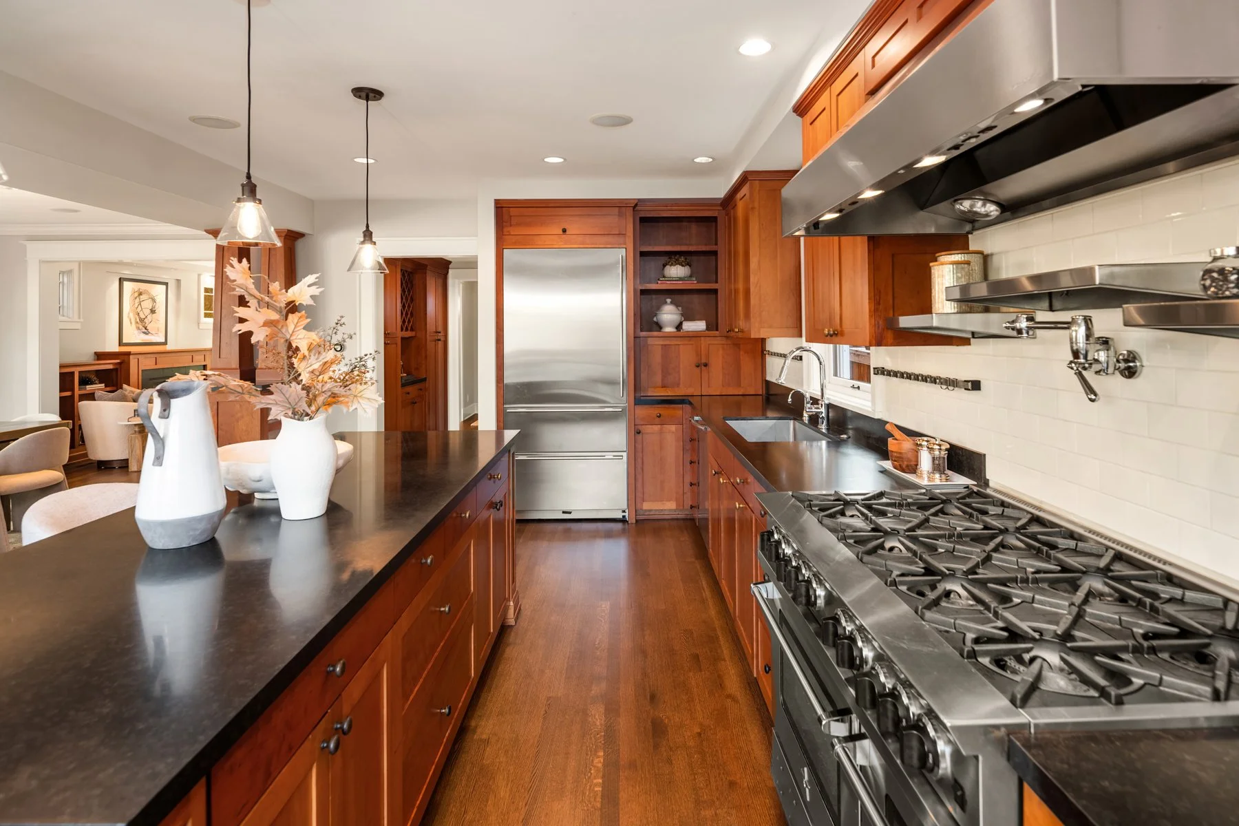 Modern kitchen with wooden cabinets, stainless steel appliances, black countertops, and white backsplash tiles. There are hanging pendant lights and decorative items on the counter.