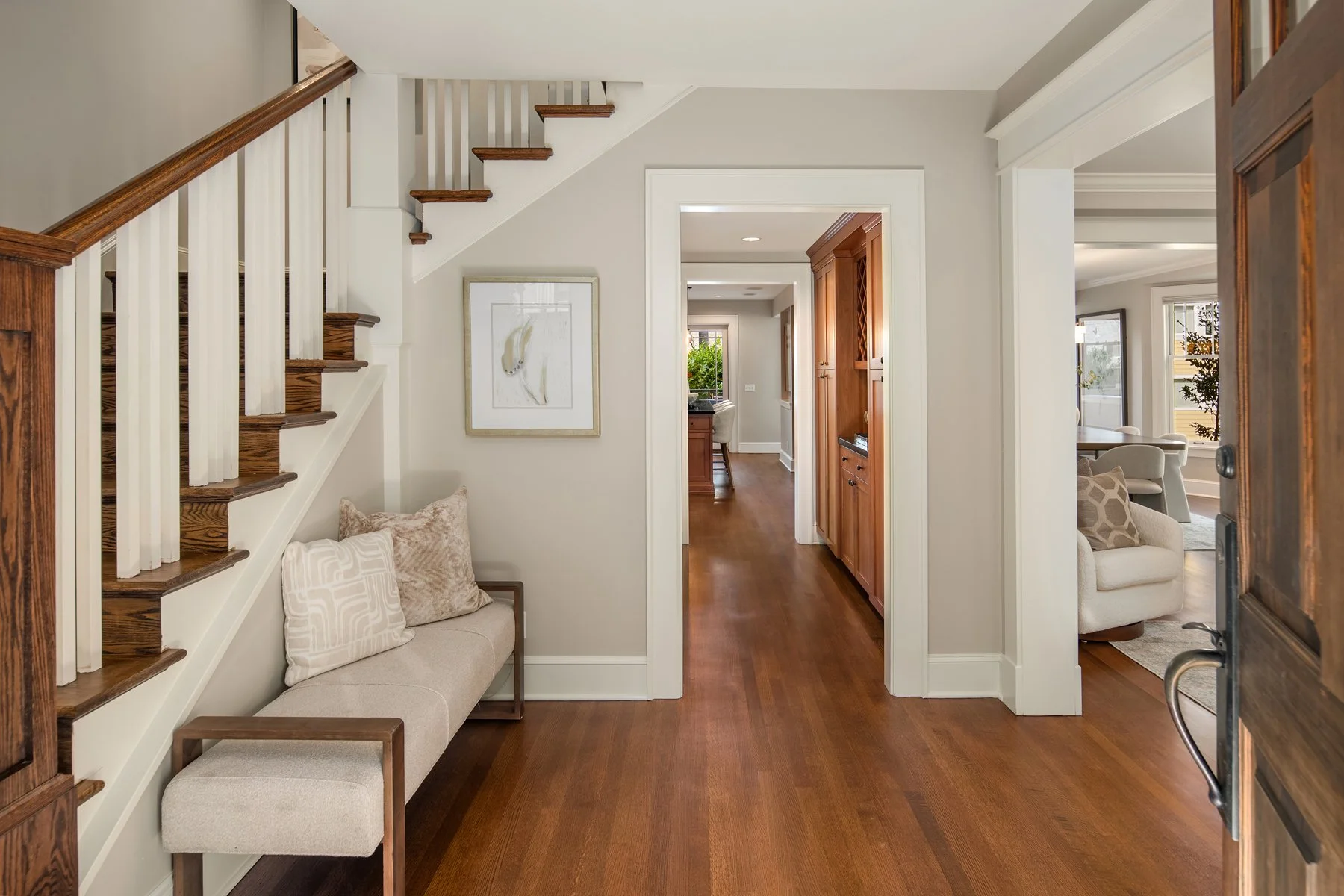 Entryway with staircase on left, seating area with pillows below stairs, hallway leading to living room and kitchen, hardwood floors, and neutral wall colors.