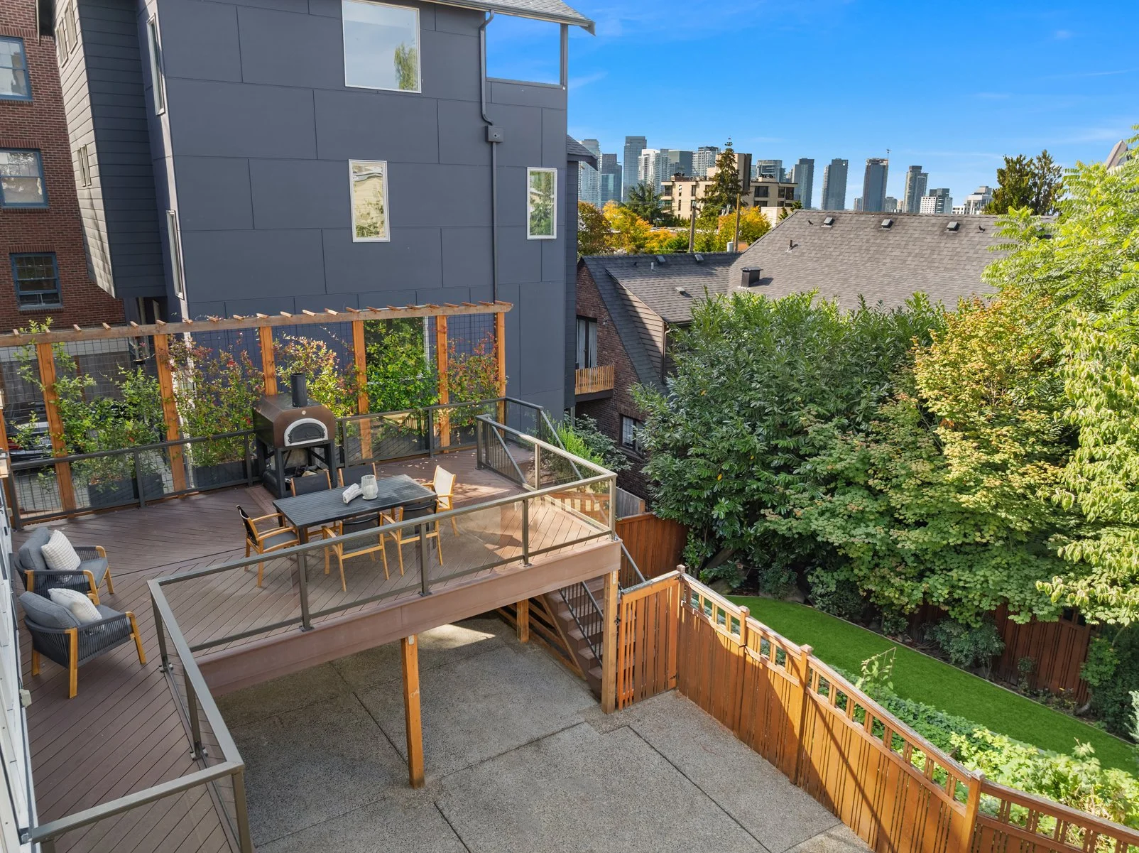 Backyard patio with wooden deck, outdoor furniture, a pizza oven, and lush trees, with city skyline in the background on a sunny day.