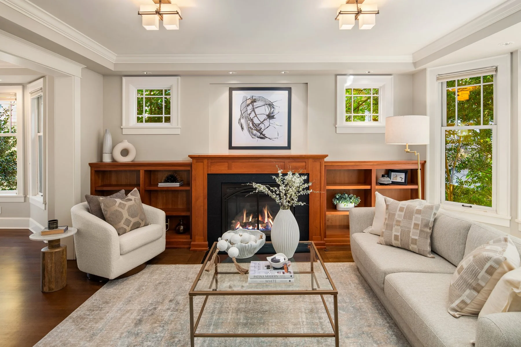 Modern living room with fireplace, beige couch, white armchair, coffee table with decorative objects, artwork above fireplace, and large windows with greenery outside.