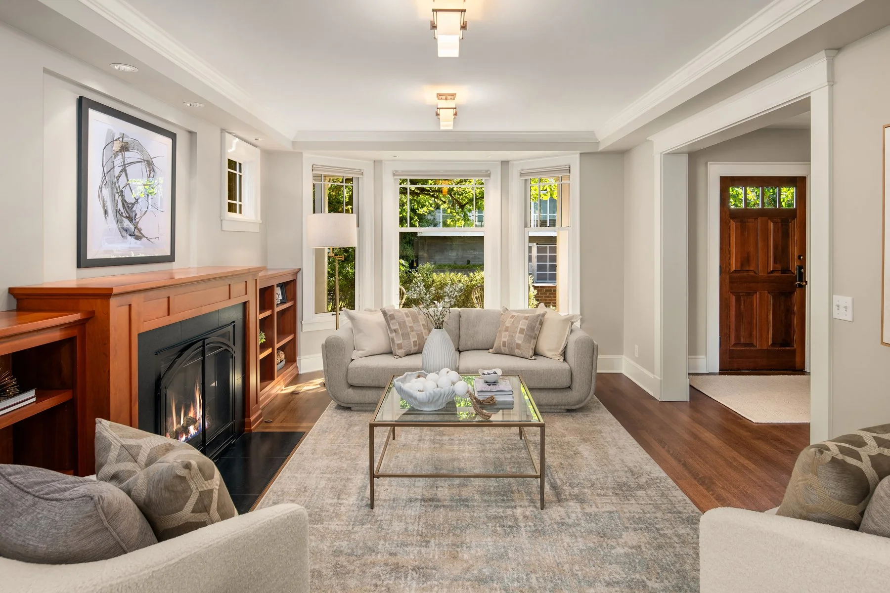Bright living room with large bay window, beige sofa with beige and patterned pillows, a glass coffee table with decorative items, a fireplace with a wooden mantel, framed abstract art, and a dark wood front door.