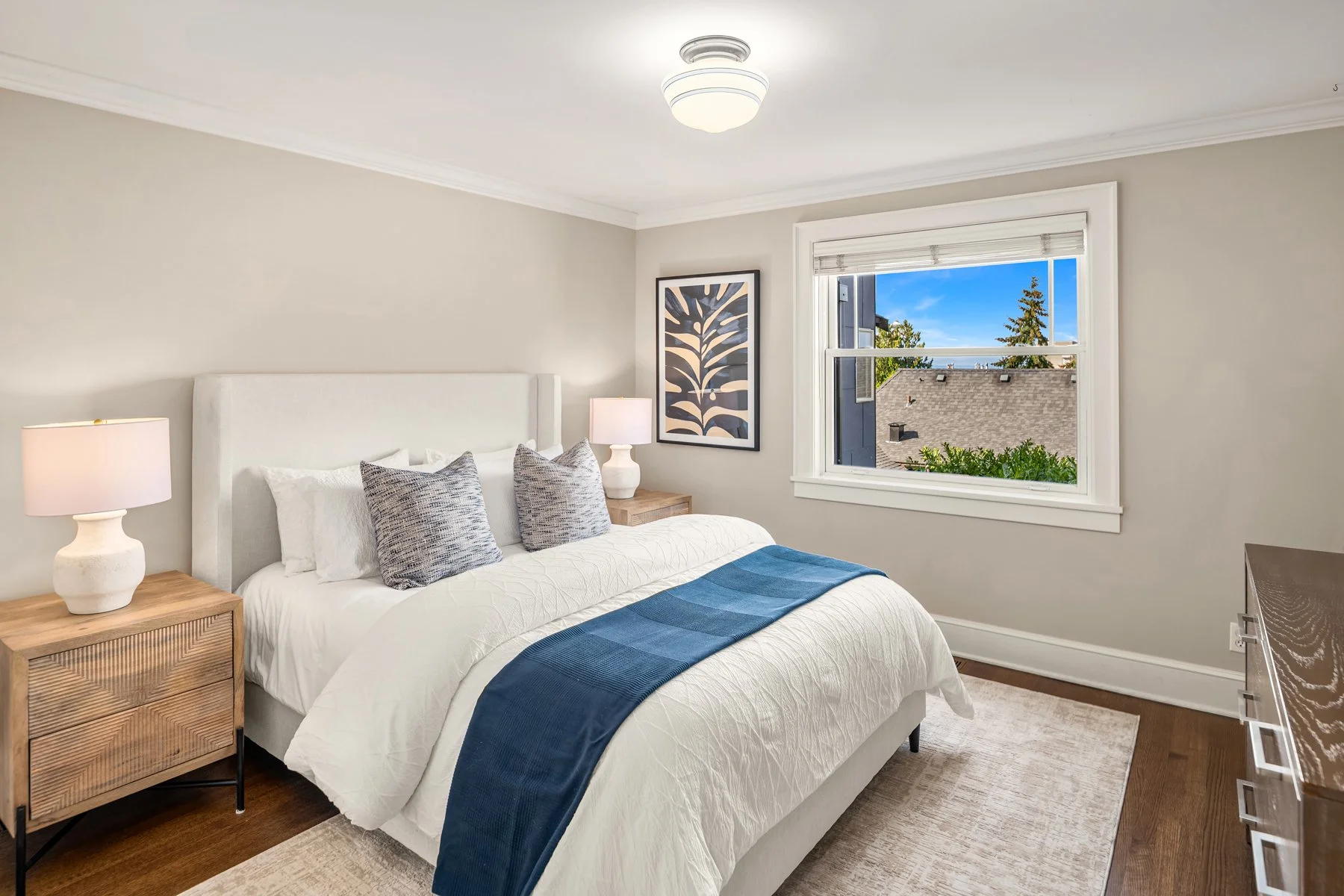 A bedroom with a large window showing a view of a roof and trees outside. The room has a white bed with a blue throw and two patterned pillows, wooden nightstands with white lamps, and a framed leaf artwork on the wall.