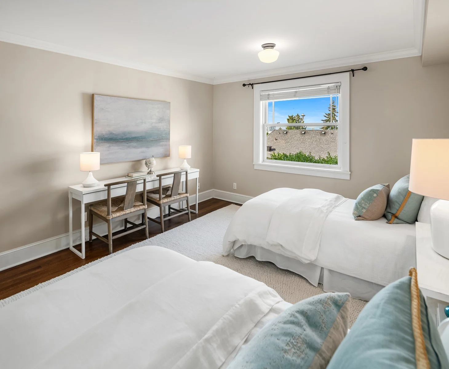 A bright, minimalist bedroom with two beds with white bedding and pastel-colored pillows, a white desk with two lamps and chairs, a large window showing rooftops and greenery outside, and a framed abstract landscape painting on the beige wall.