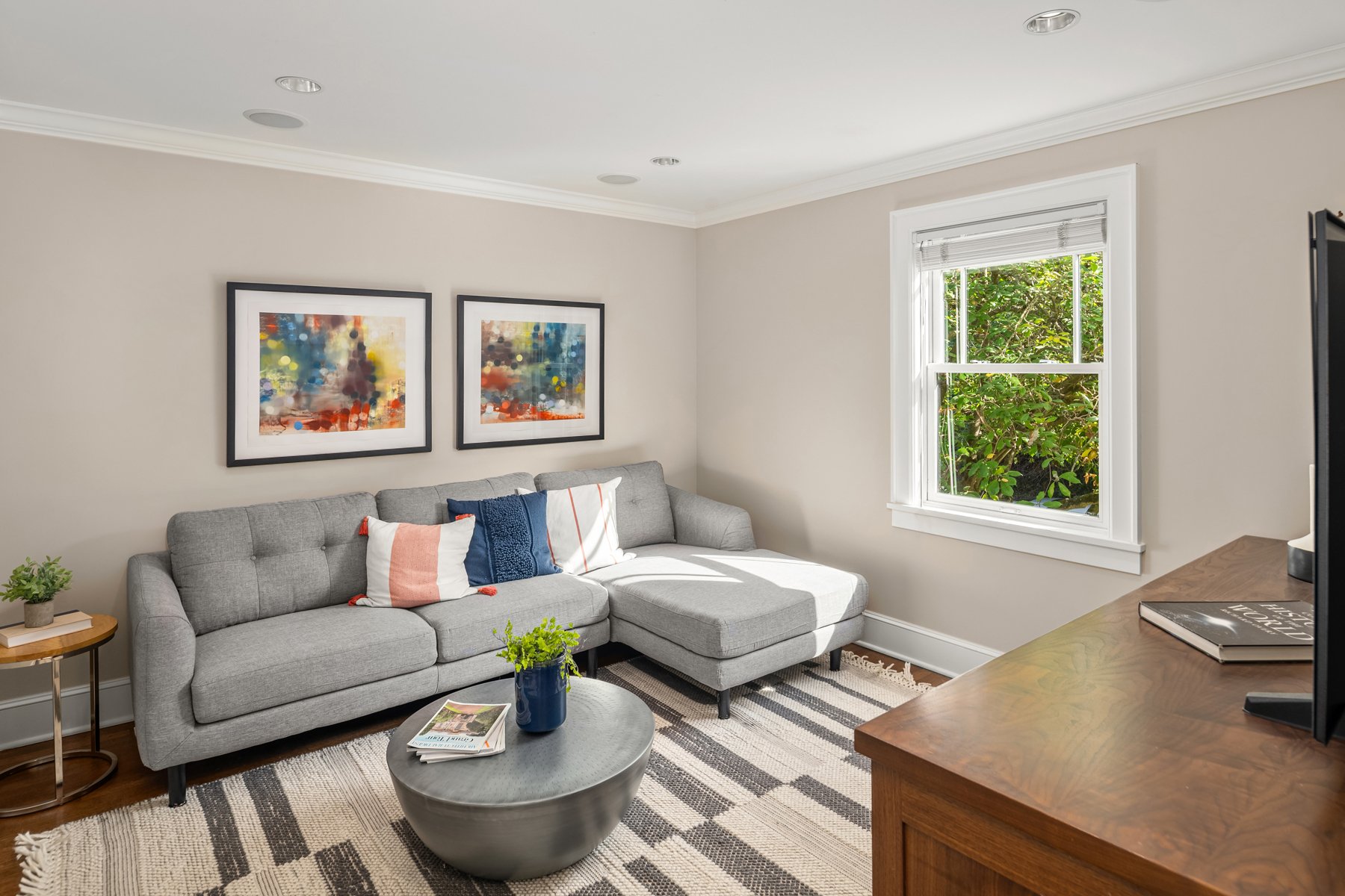 Living room with a gray sectional sofa, striped rug, wall art, a coffee table with magazines, and a window with greenery outside.
