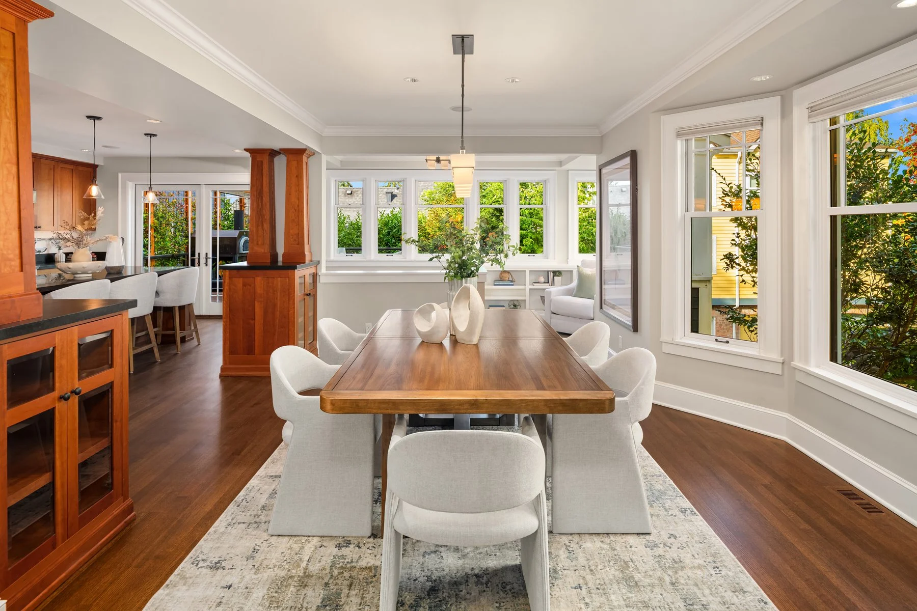 Bright, open dining and living area with large windows showing greenery outside. Wooden dining table surrounded by white upholstered chairs, decorative vases on the table, and a chandelier hanging overhead. Kitchen area with wooden cabinets and bar s