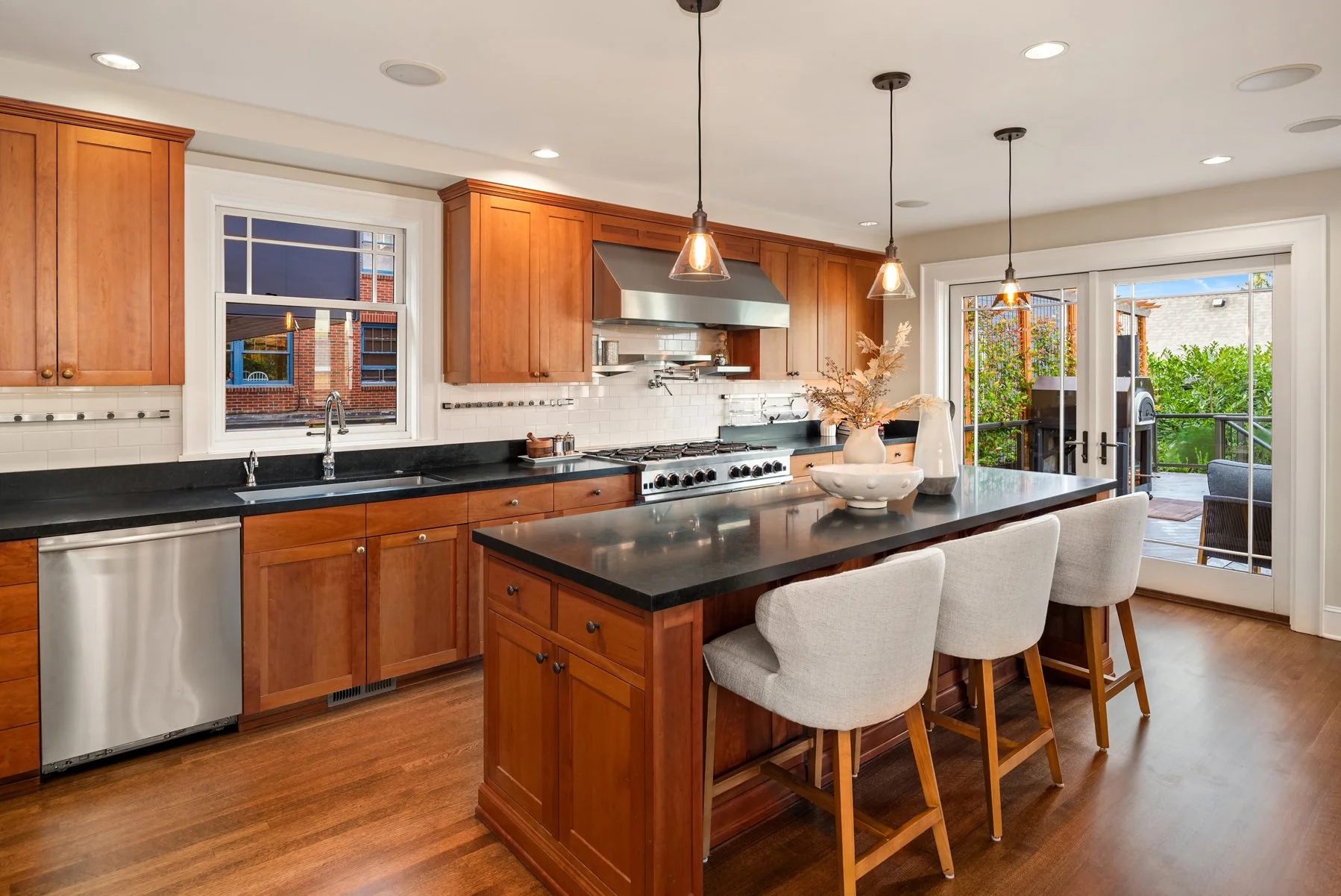 Modern kitchen with wooden cabinets, black countertops, stainless steel appliances, and a breakfast bar with three cream-colored chairs. Large window and sliding glass door overlook an outdoor patio with greenery.