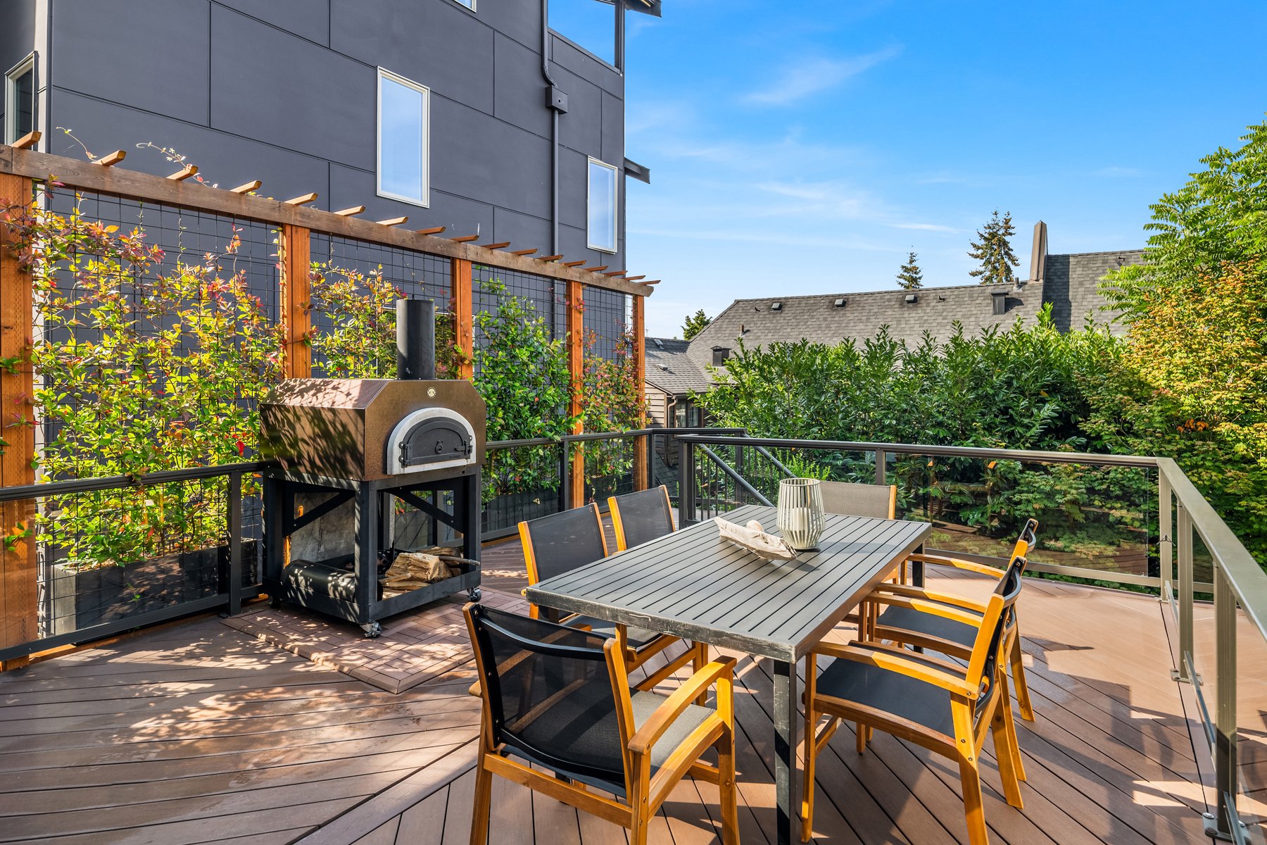 Outdoor balcony with a dining table, chairs, a pizza oven, surrounded by greenery and modern buildings under a clear blue sky.