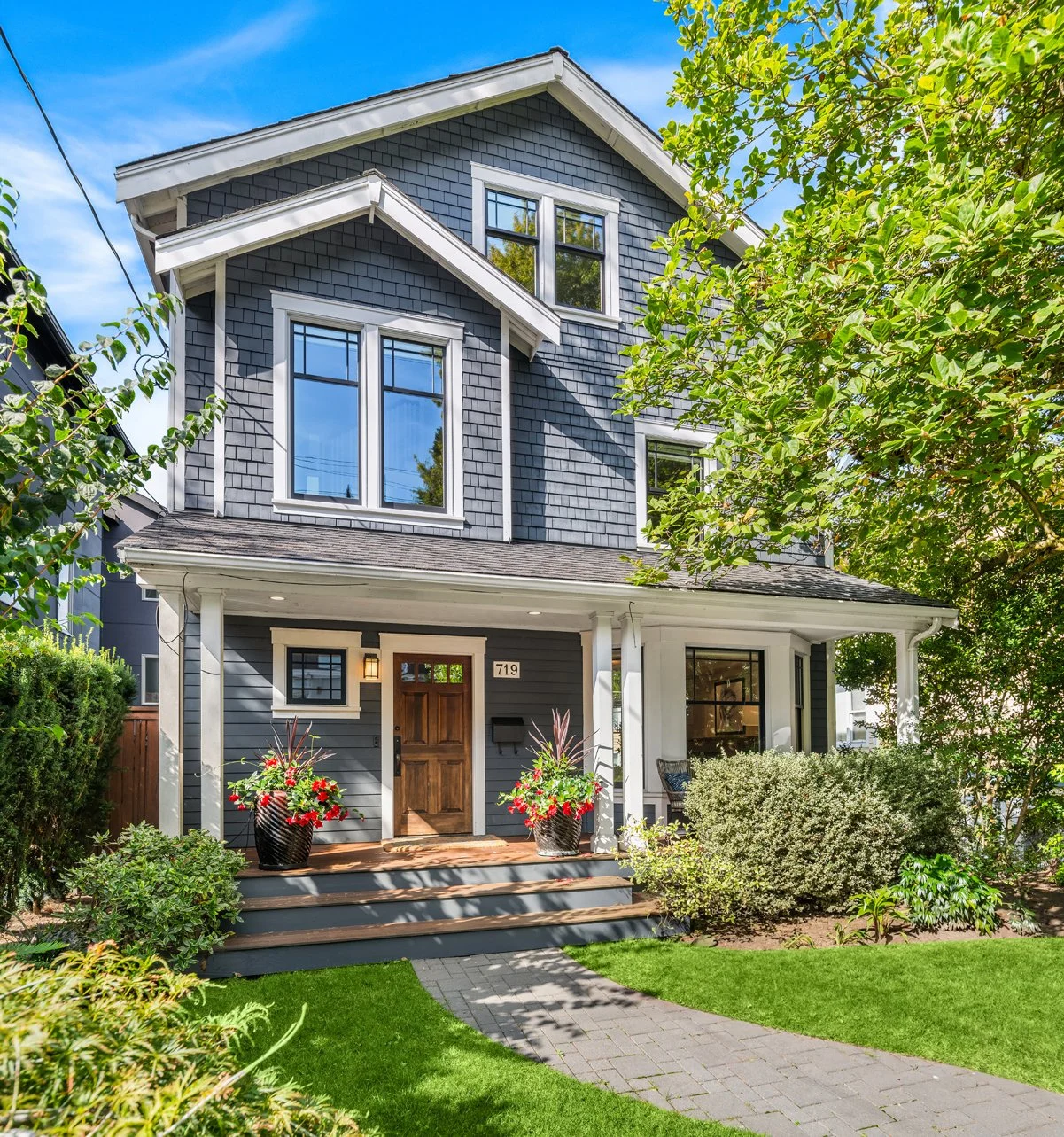 Beautiful three-story house with gray siding, white trim, and a wooden front door. The house has large windows, a covered porch with potted plants, and a well-maintained front yard with lush green grass, shrubs, and trees. A brick pathway leads to th