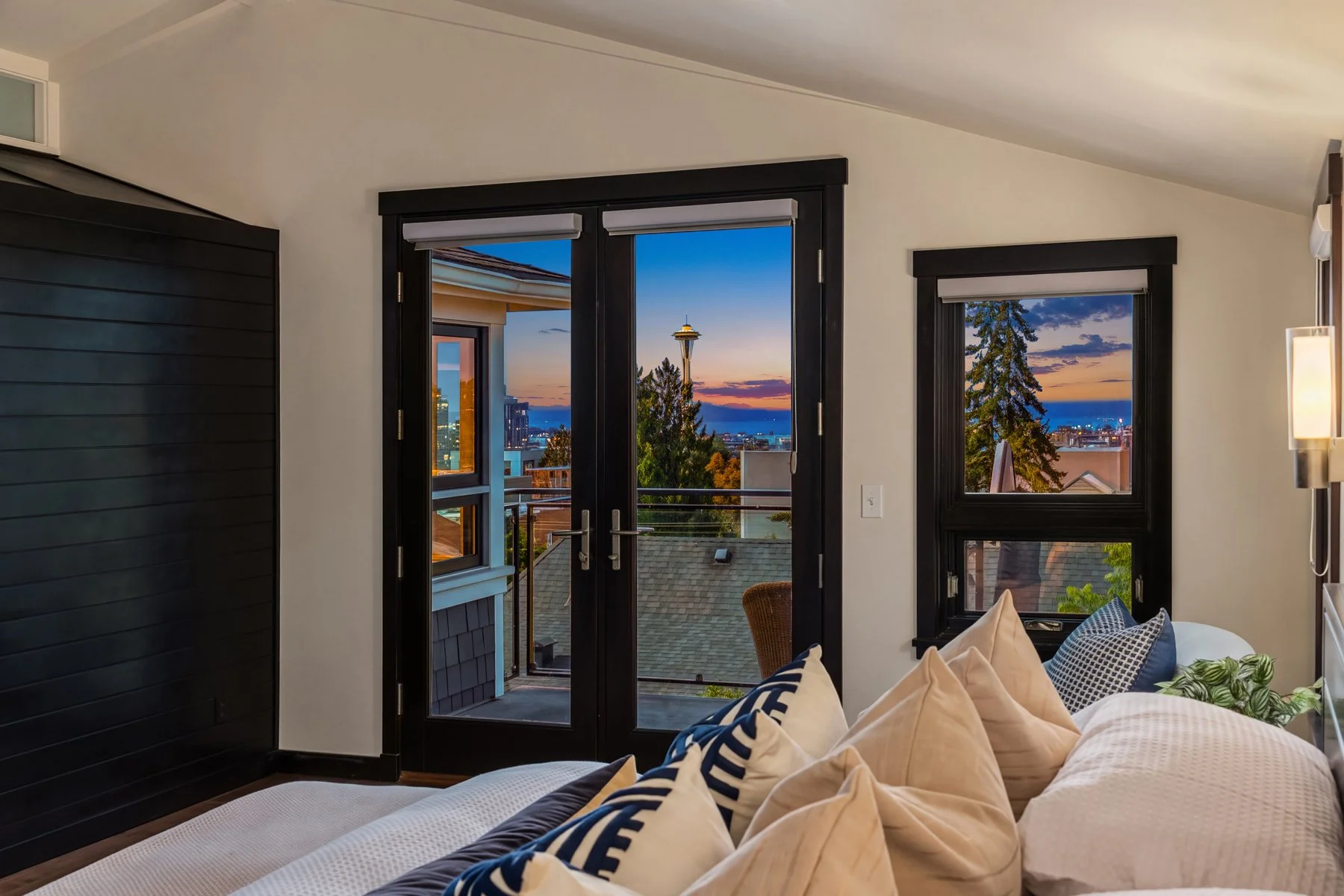 Interior of a modern bedroom with large windows and a balcony door showing a city view with the Space Needle at sunset. The room has black window frames, white walls, and a bed with beige, navy, and patterned pillows.