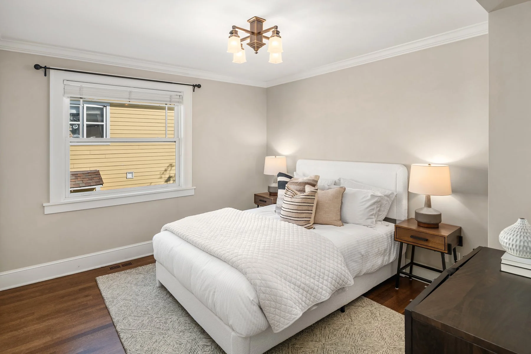 A cozy bedroom with a white bed, beige walls, and wooden nightstands with lamps. A window with blinds reveals the exterior of a house with yellow siding.