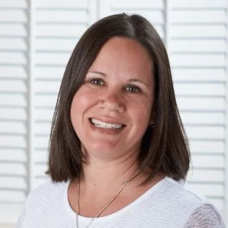 Smiling woman with shoulder-length dark hair, wearing a white top, in front of white blinds.