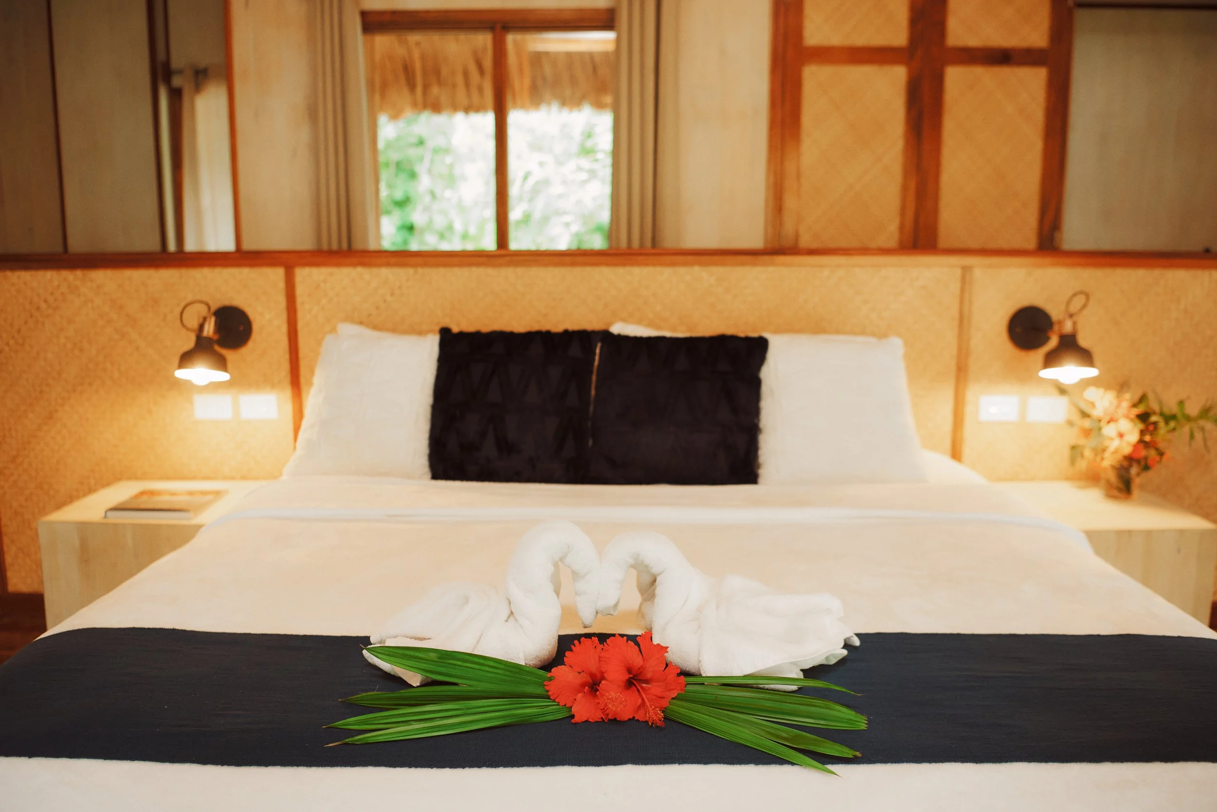 Bed made up in white with a dark headboard, two towel swans and a red flower, in a wood-walled room with side lamps