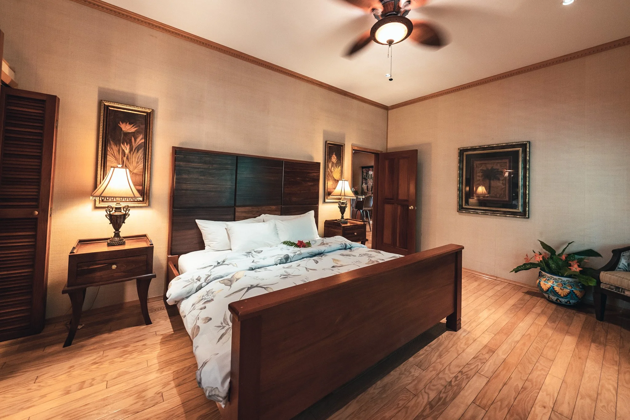 Bedroom with a wooden bed and floral bedding, matching bedside lamps, framed art, a ceiling fan and a large potted plant