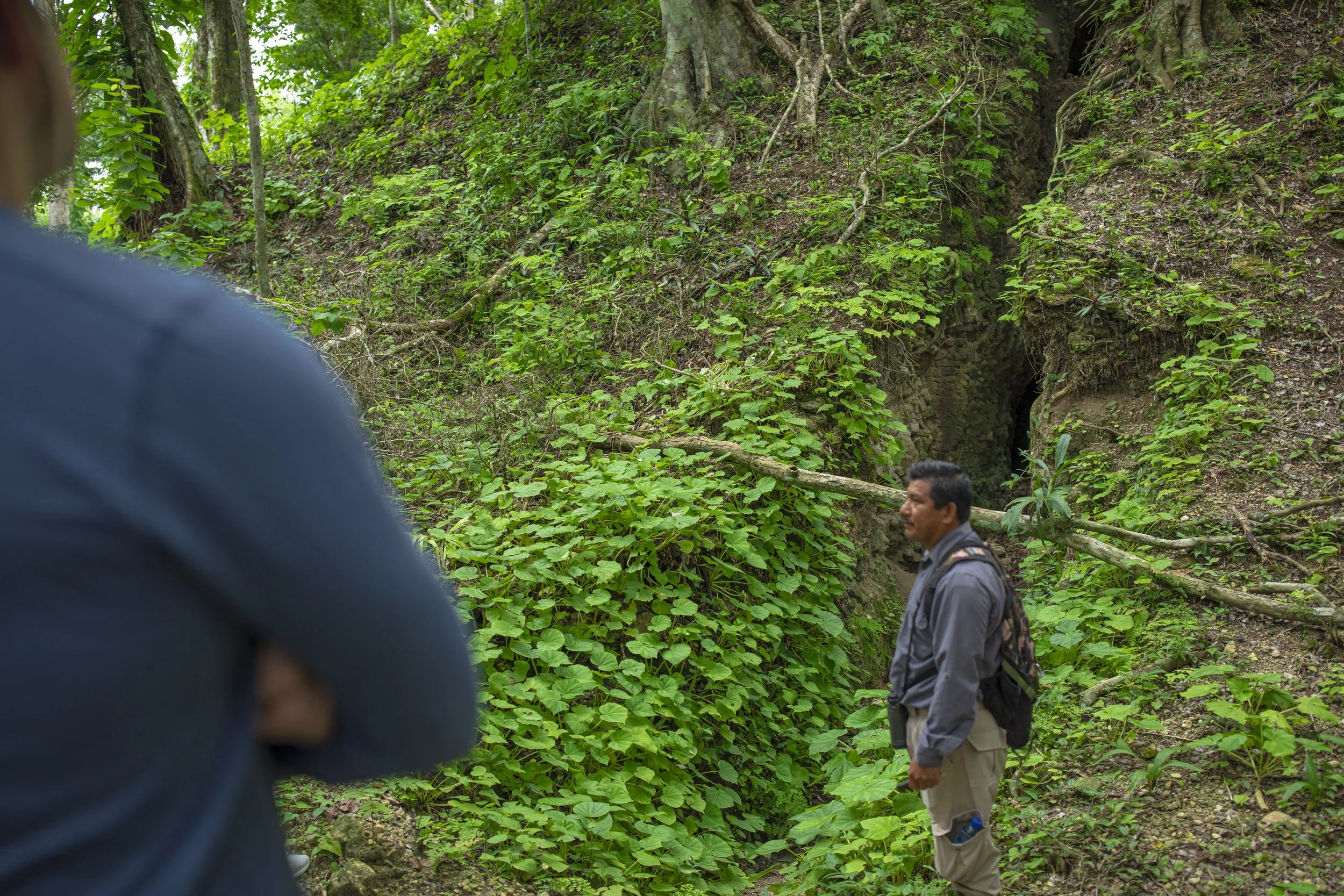 A person standing in a lush, green forest near a small cave or opening in the hillside, with another person partially visible in the foreground.